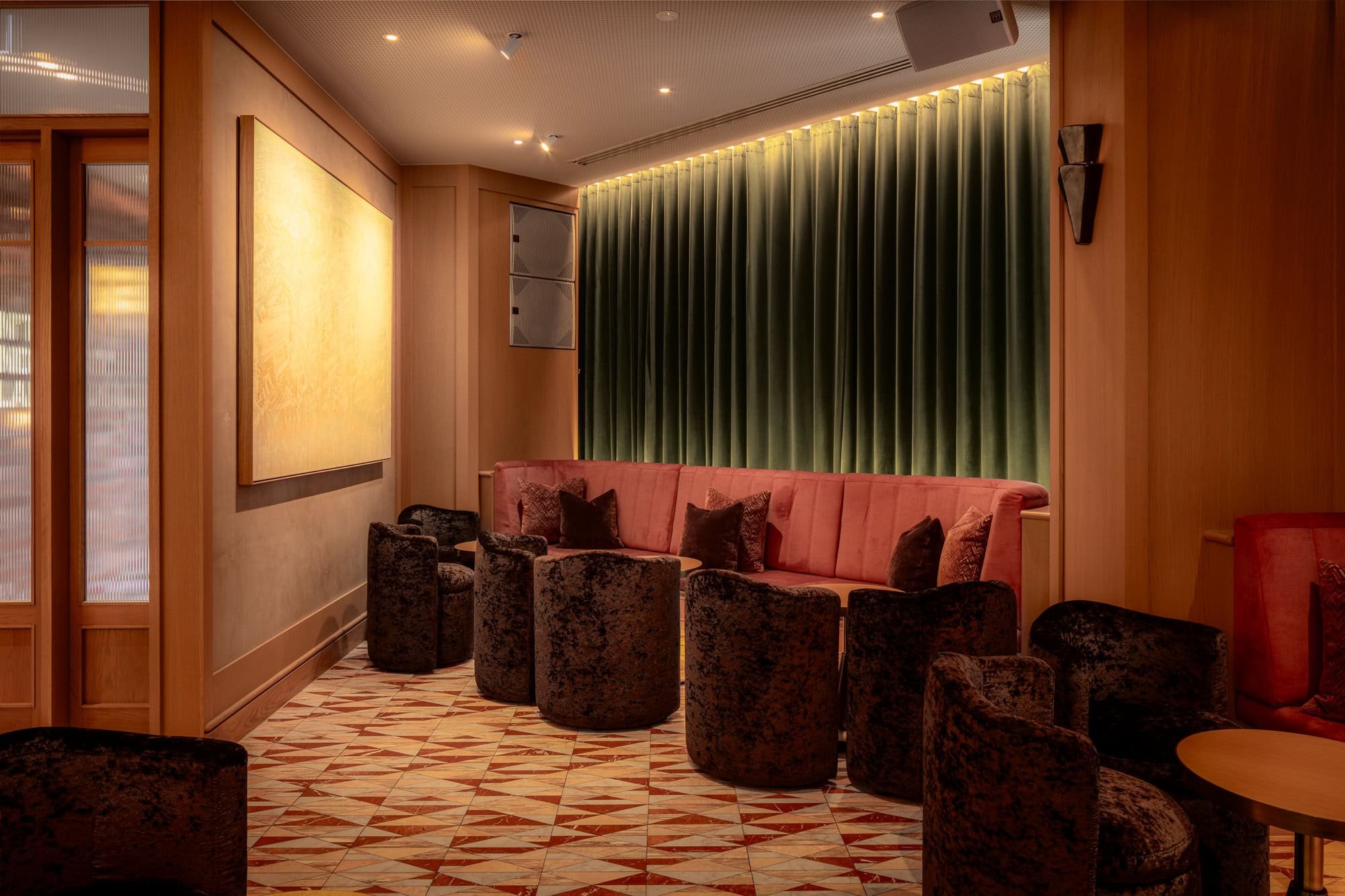 A cozy lounge area with a pink sofa and patterned armchairs arranged around small round tables, a green velvet curtain on the wall, warm lighting, wooden paneling on the walls, and a patterned tile floor.