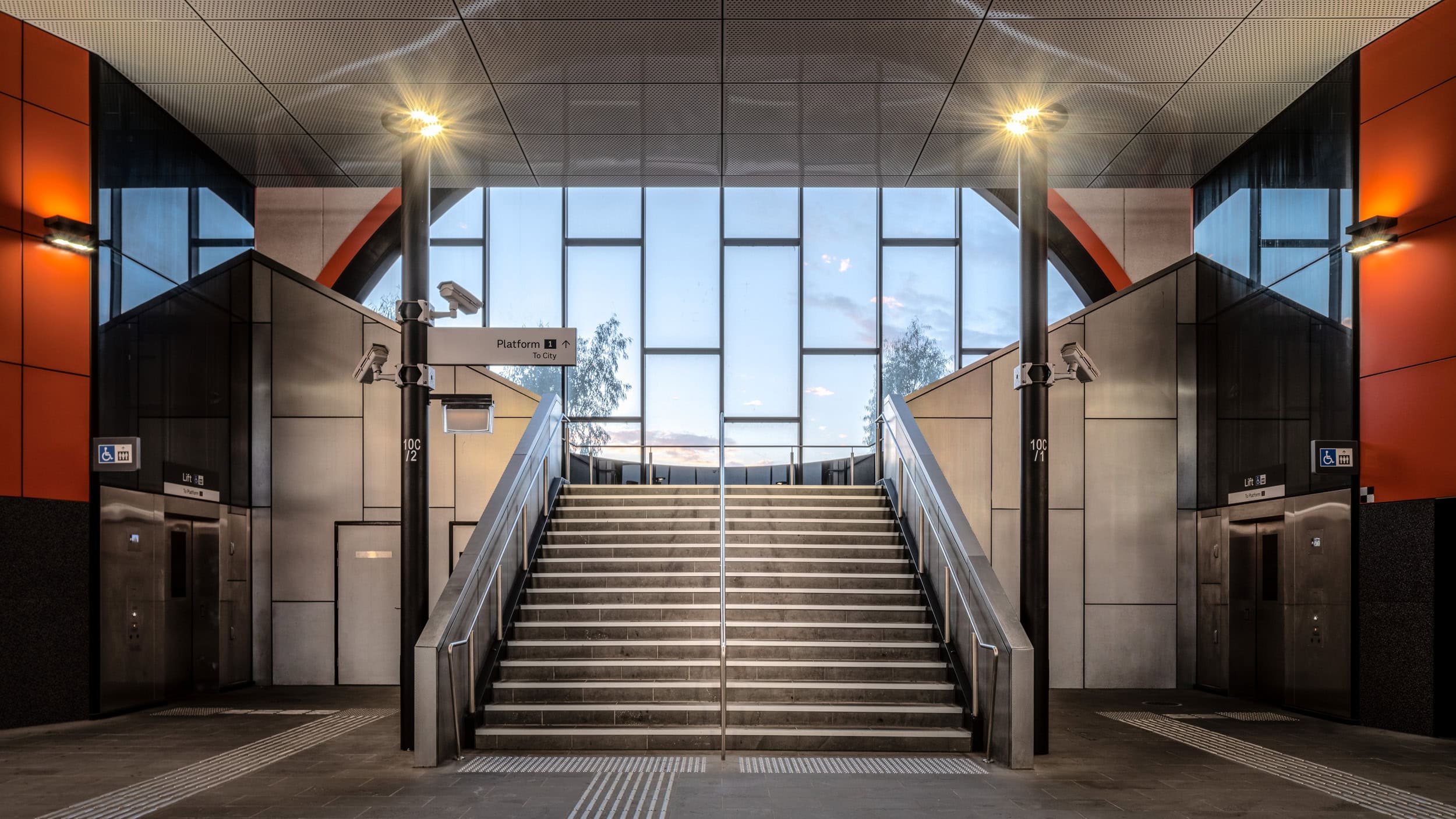Stairs leading to an upper level in a modern transportation station, with large windows, escalators on sides, safety signs, and surveillance cameras.