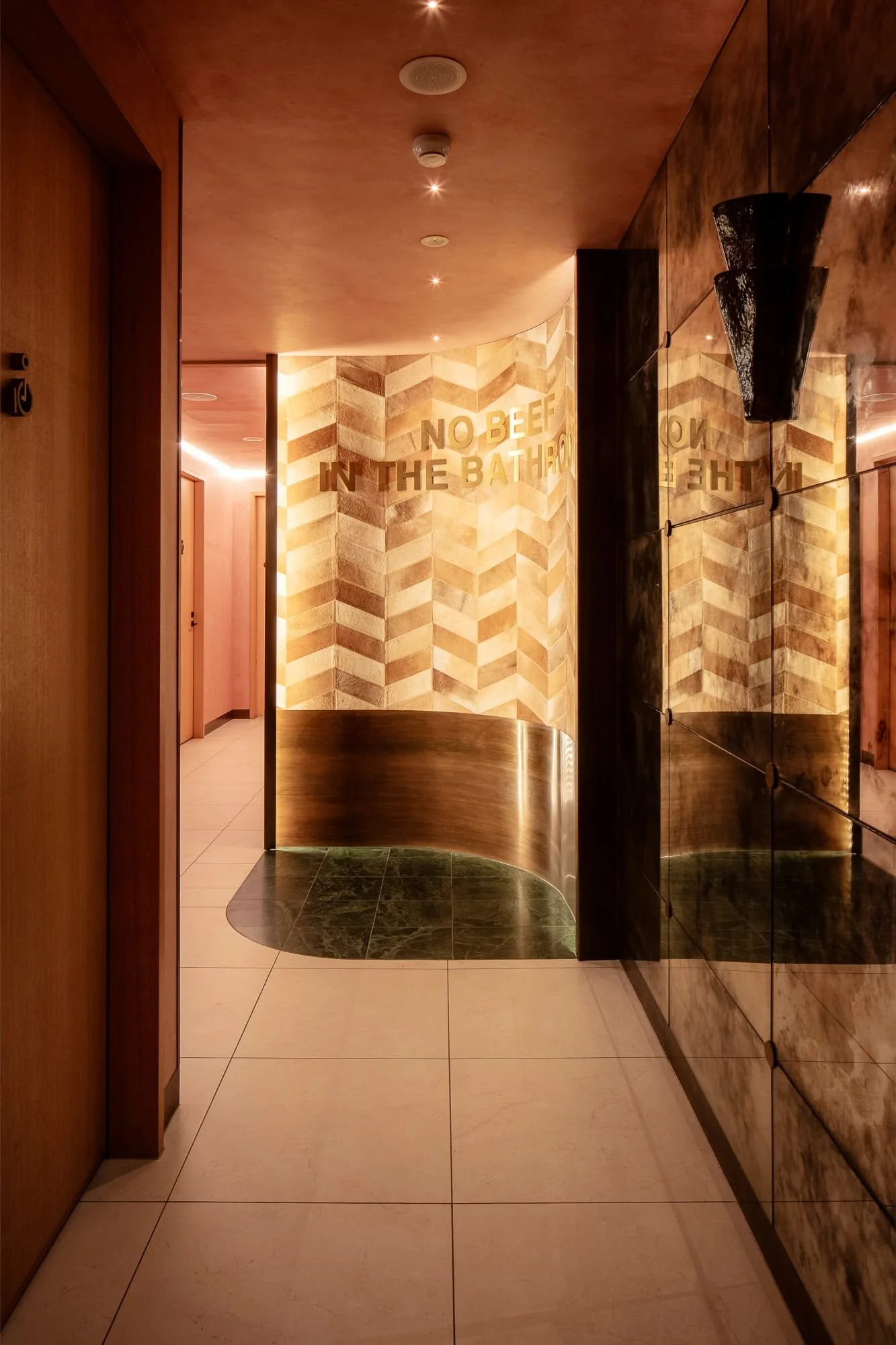 Hallway in a modern building with a curved wall and warm lighting. There's a sign that says 'No Beef in the Bathroom' on a stone textured wall. The floor has white and dark green tiles, and the ceiling has small, round lights.