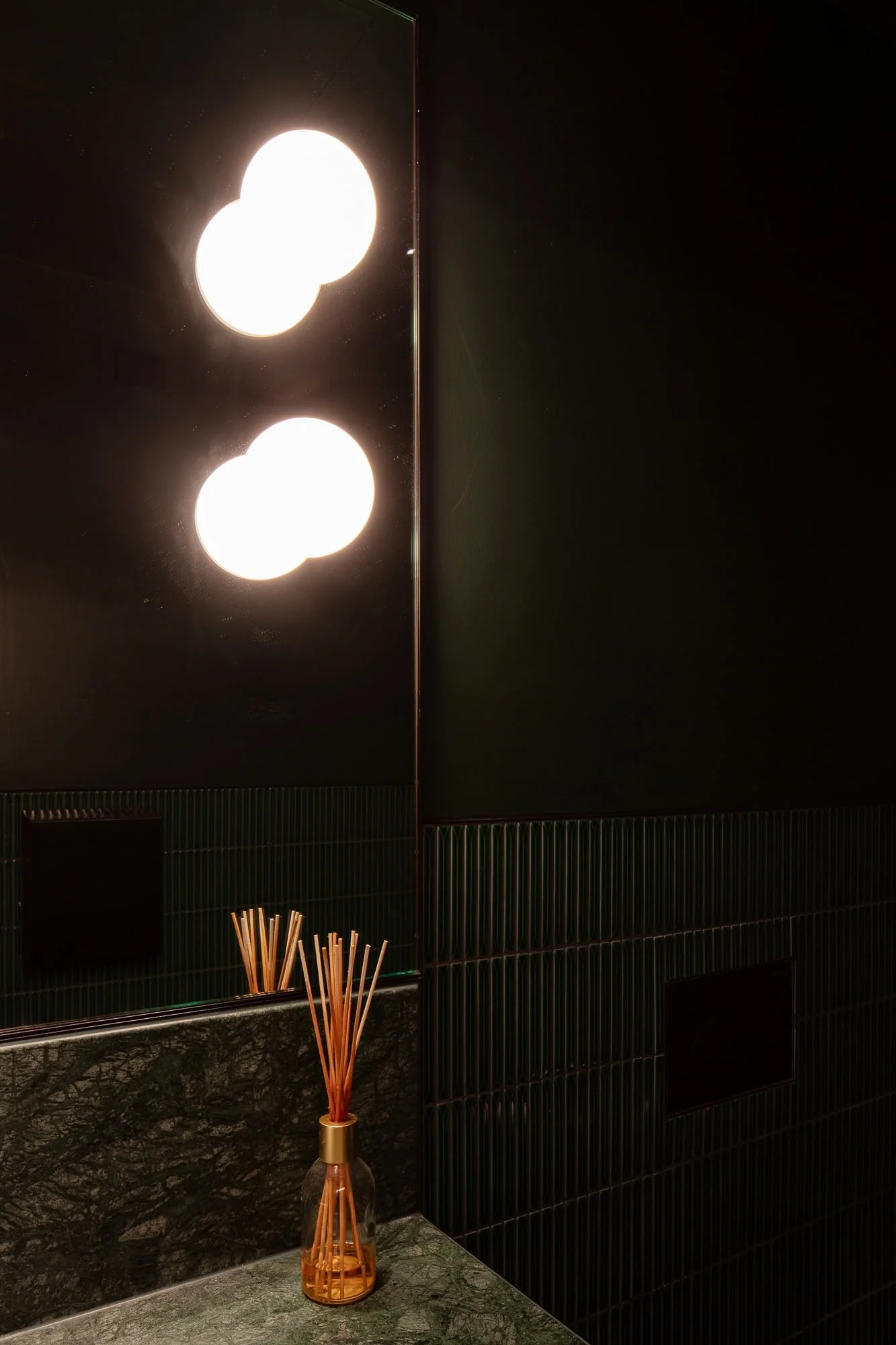 A room with black walls and ceiling, two bright ceiling lights, a green marble countertop, and a small glass vase with brown reed diffuser sticks placed on the countertop.
