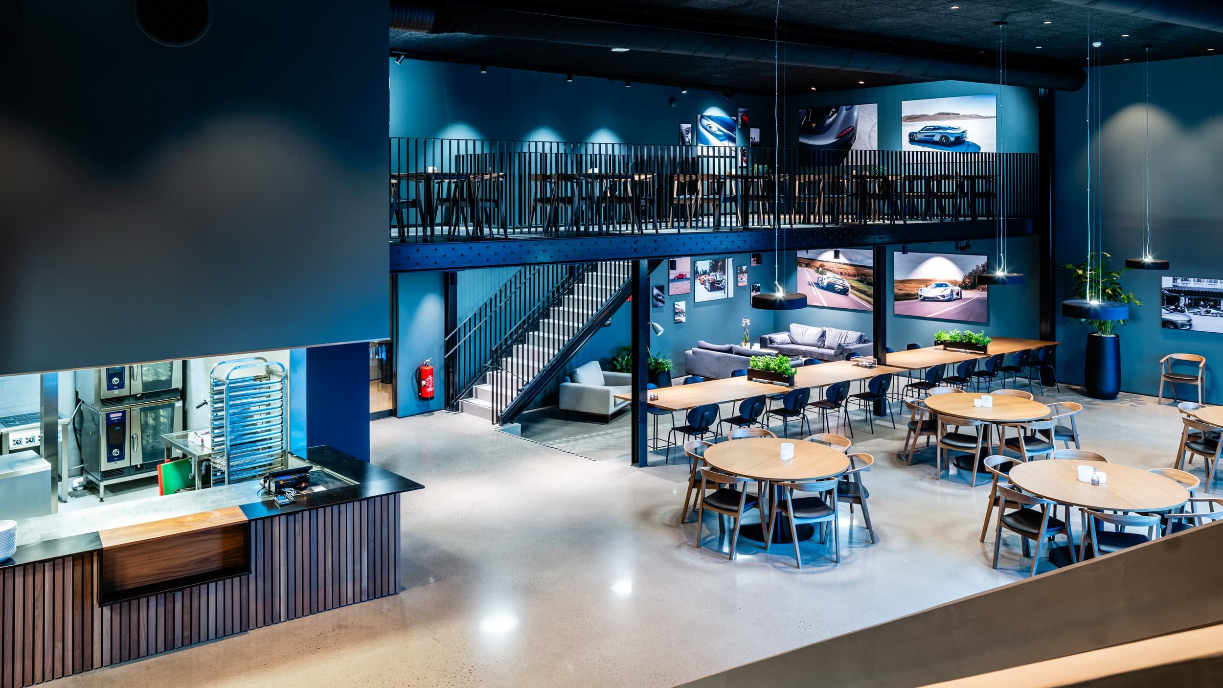 Modern car showroom or lounge area with multiple tables, chairs, blue walls, framed car photos, hanging lights, and an upstairs balcony with additional seating.