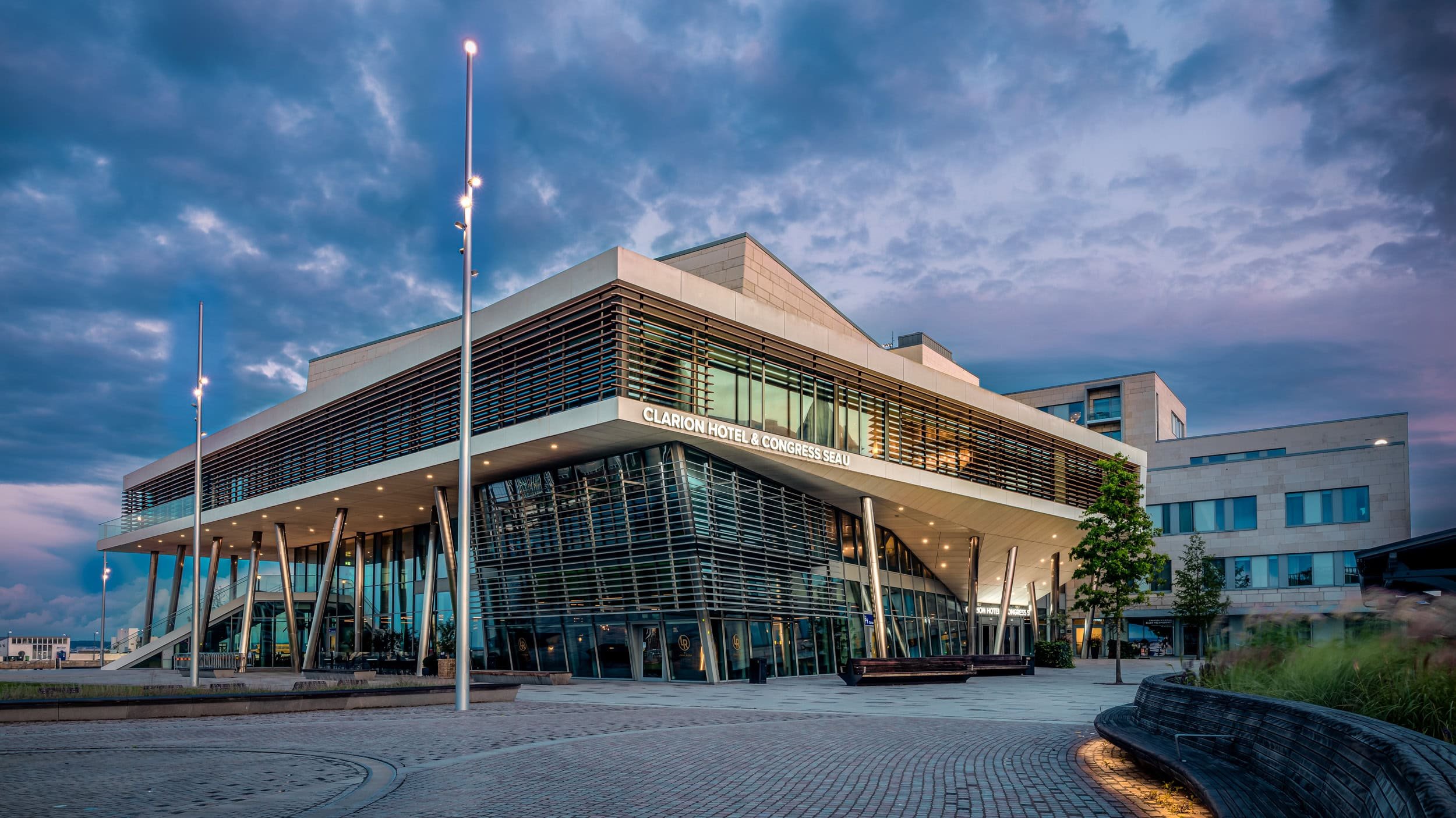 Exterior view of the Clarion Hotel & Congress Seau with modern architecture, glass walls, and a landscaped plaza at dusk.