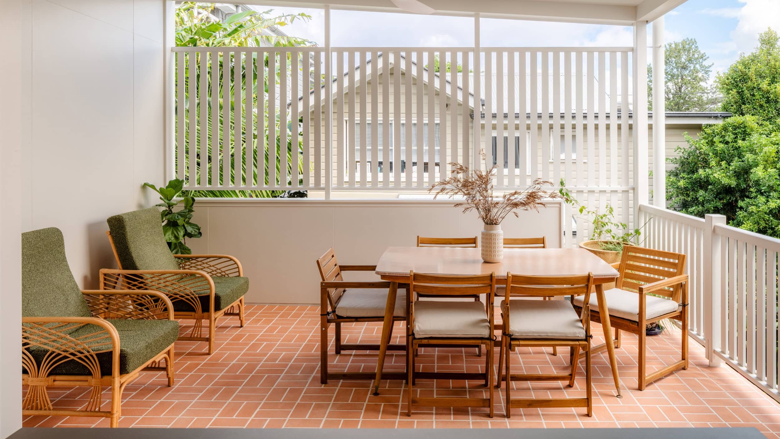 An outdoor porch with a wooden dining table, six chairs, green cushioned chairs, and potted plants, overlooking neighboring houses and trees.