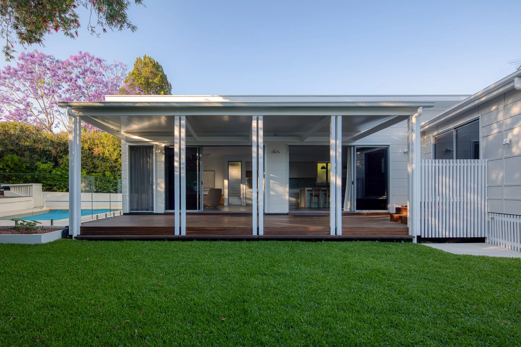 Rear view of a modern house with a wooden deck and a sliding glass door, surrounded by a green lawn and flowering trees, under a clear blue sky.