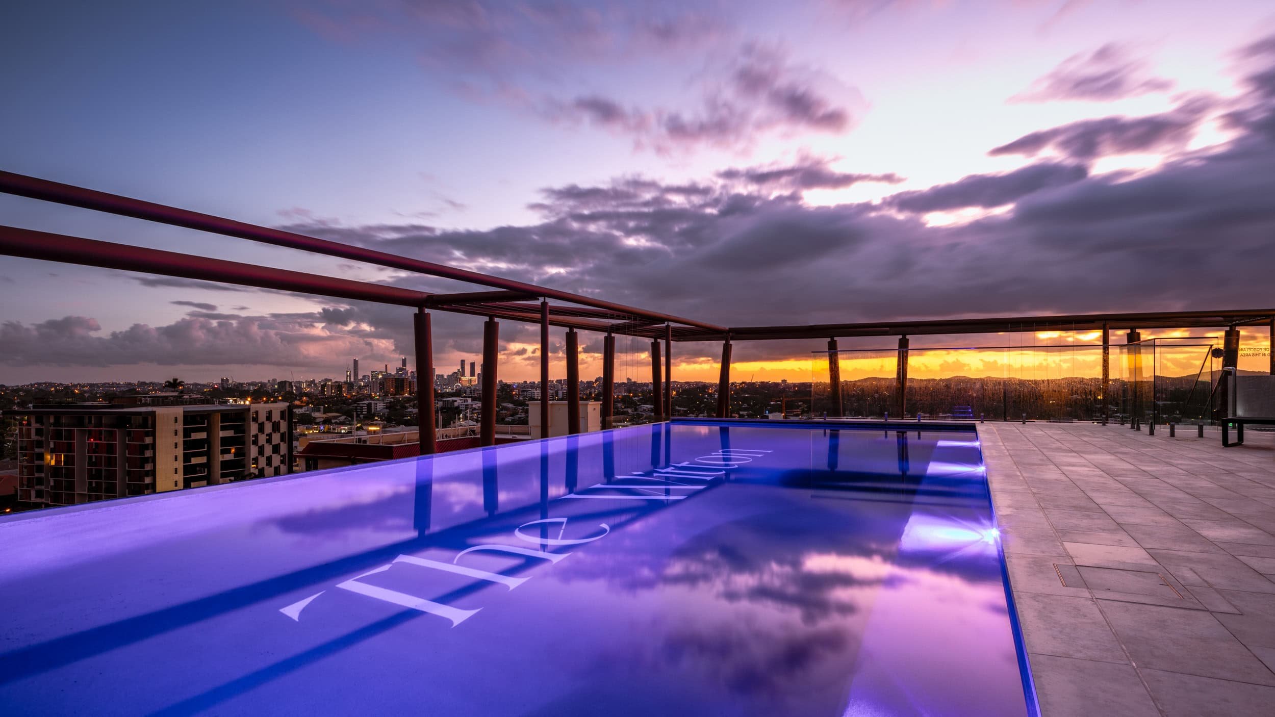 Rooftop swimming pool with a view of a city skyline at sunset, illuminated with blue lights and transparent glass barriers.