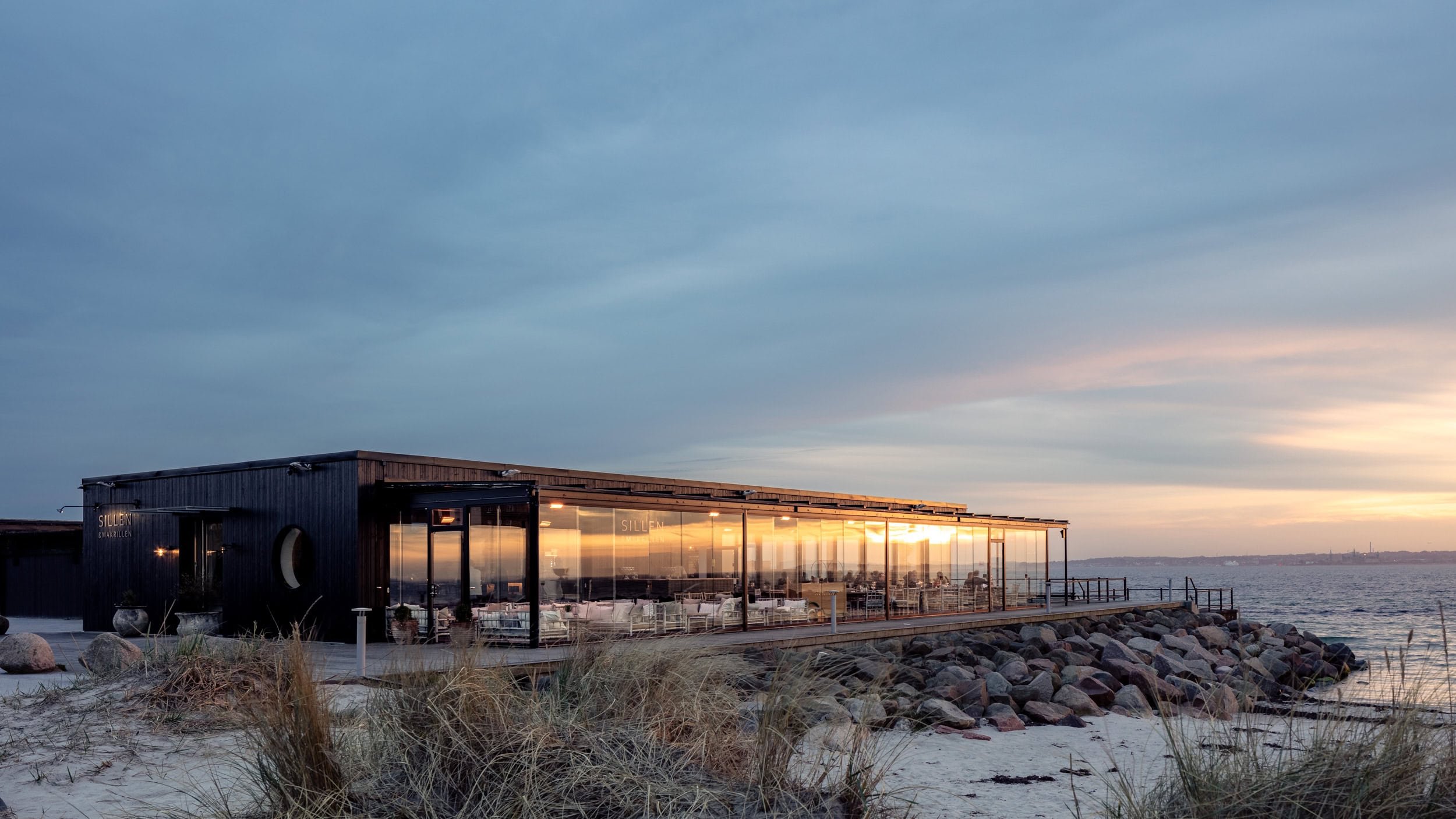 Modern black building with large glass windows overlooking the water, with sunset reflections, rocks, and beach grass in the foreground.