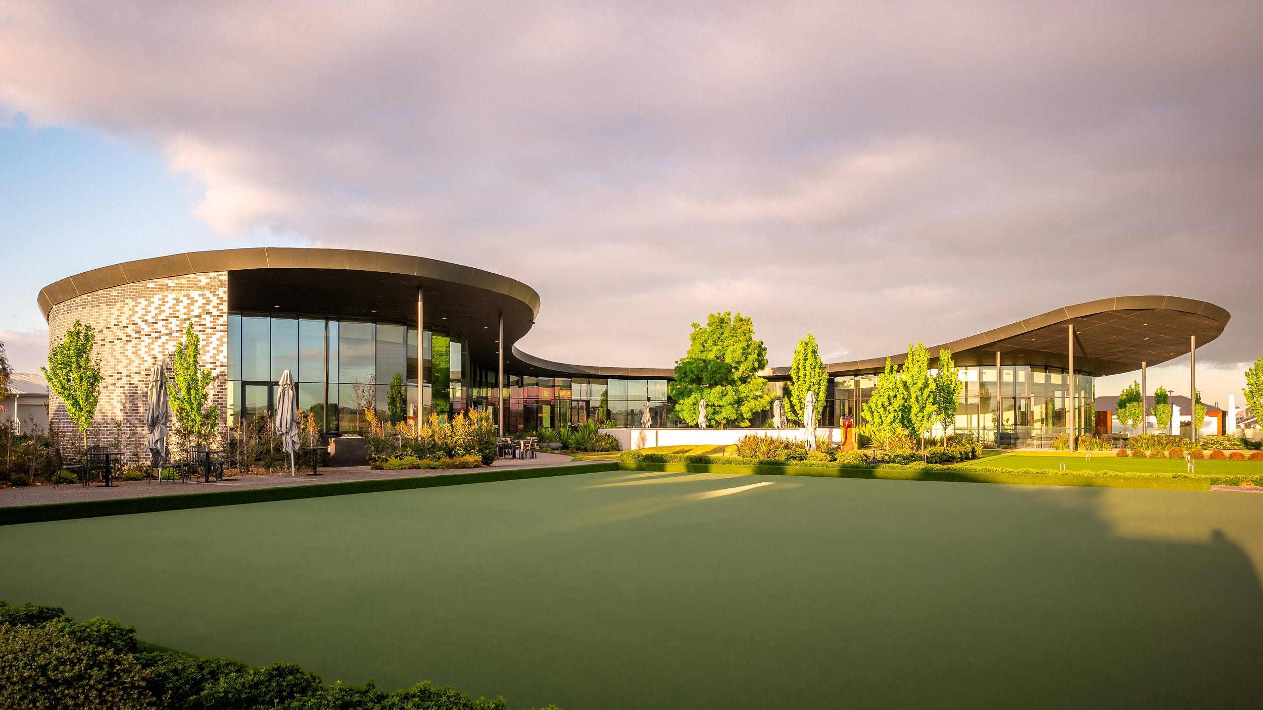 Modern building with large glass windows and curved roof, surrounded by trees, shrubs, and a manicured lawn, under a partly cloudy sky.
