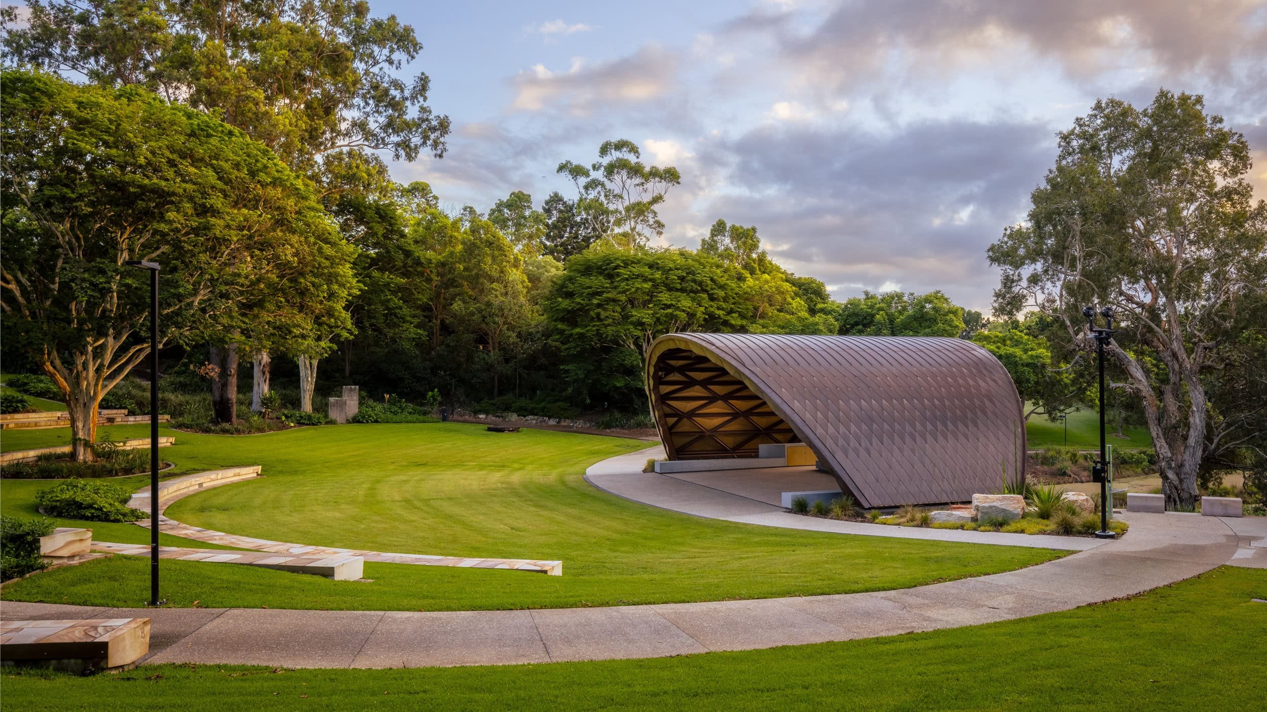 A park with a curved pavilion, lush green grass, mature trees, benches, and path lights during late afternoon or early evening.