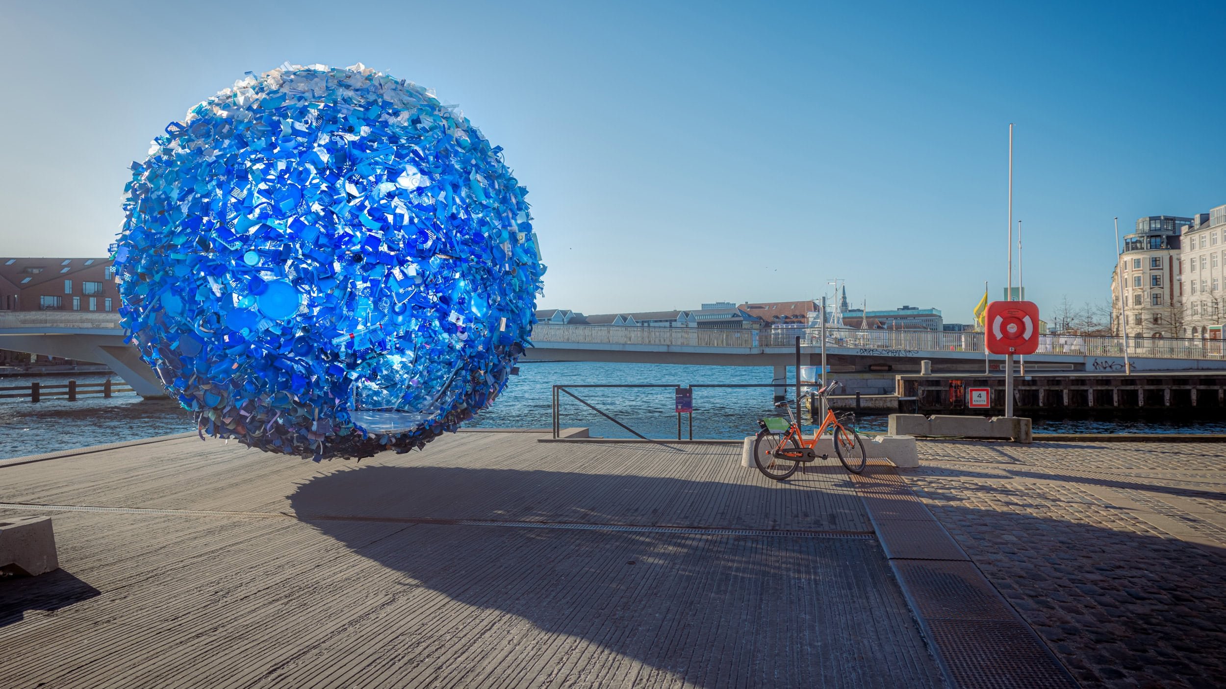 Large spherical sculpture made of blue recycled materials, situated on a waterfront promenade, with bicycle and buildings in the background.