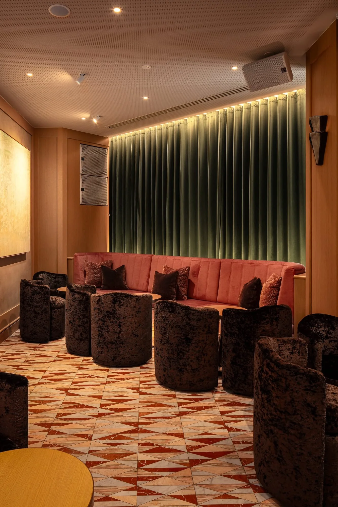 Interior of a lounge or waiting area with a pink sofa surrounded by black velvet chairs, green curtains, a patterned tiled floor, and wooden wall panels.
