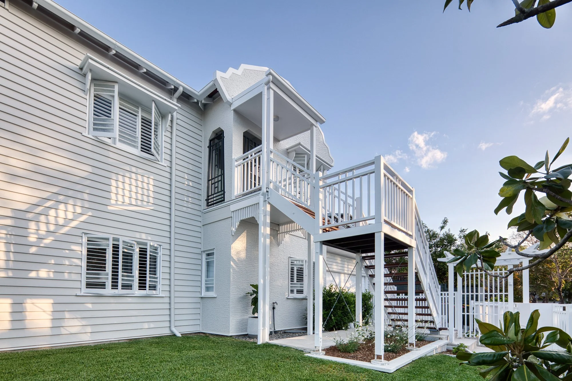 Exterior view of a white house with balconies, multiple windows with white shutters, and a landscaped backyard with green grass and plants. 
