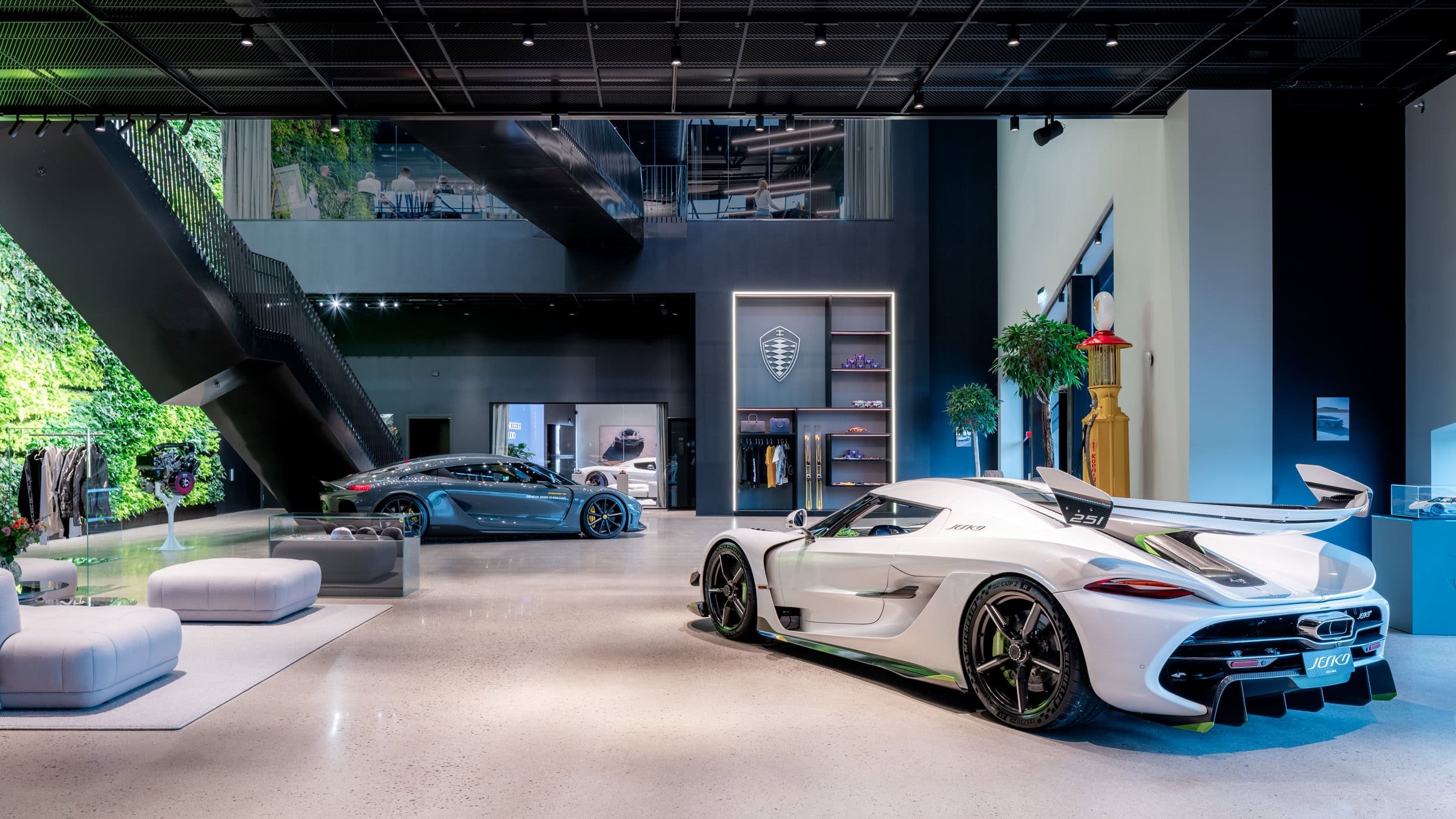 Luxury showroom displaying two sports cars, a white one in the foreground and a gray one nearby, with modern decor, greenery outside, and a staircase leading to an upper level.