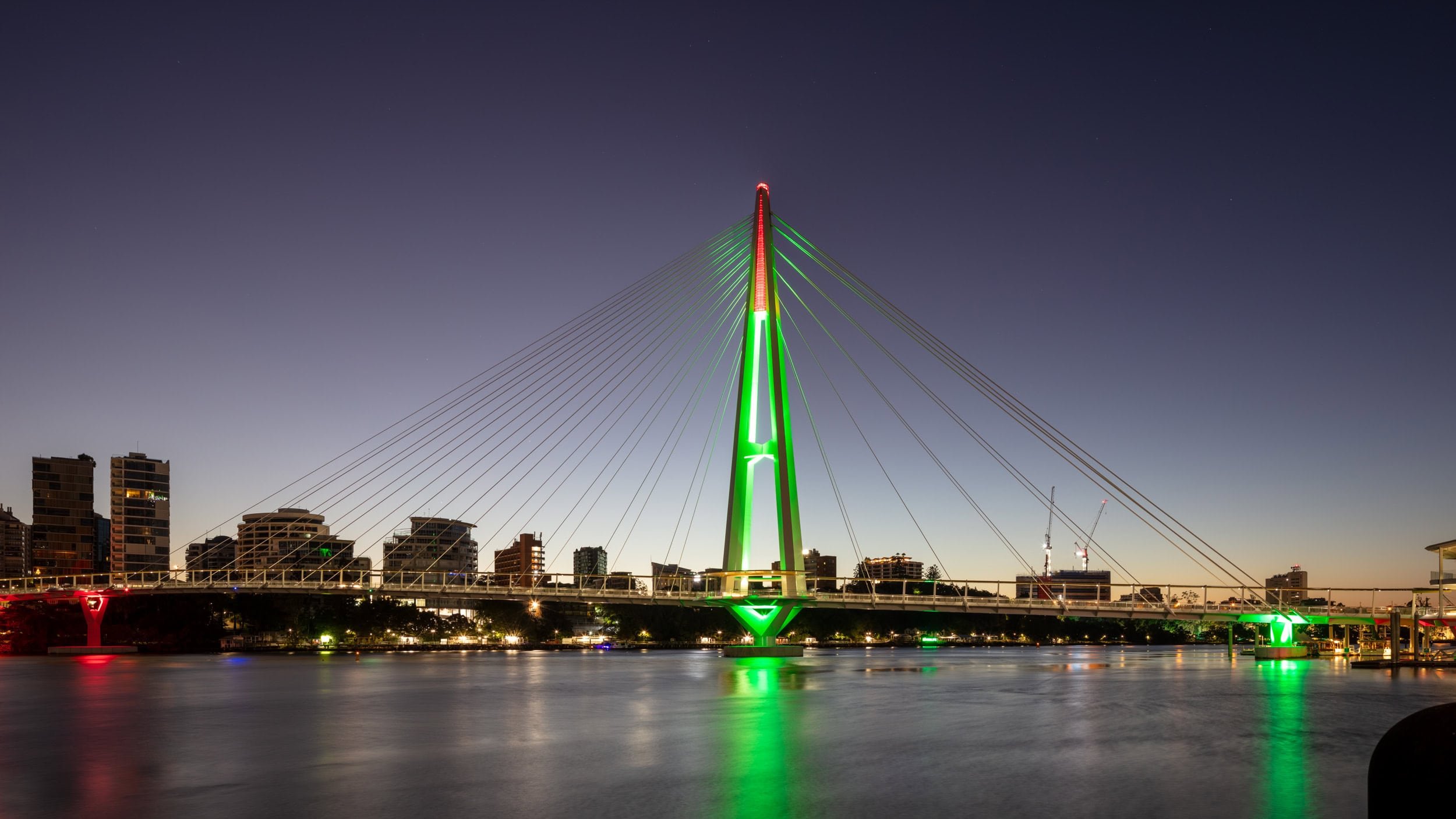 Nighttime view of a modern cable-stayed bridge illuminated with green and red lights, spanning over a body of water with a city skyline in the background.