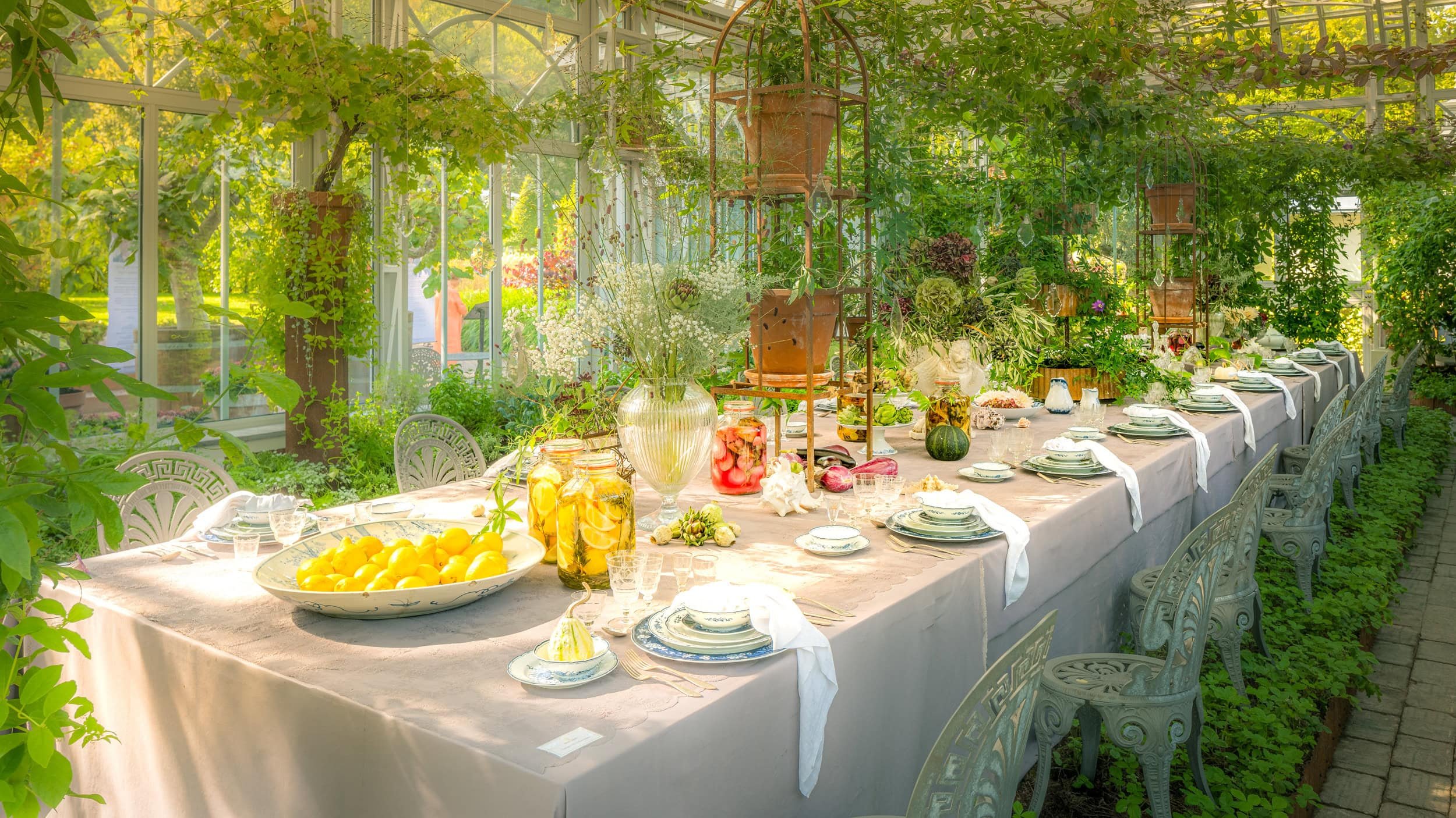 Long table set for a meal in a lush greenhouse decorated with plants, flowers, and glass jars of preserved fruits and vegetables.