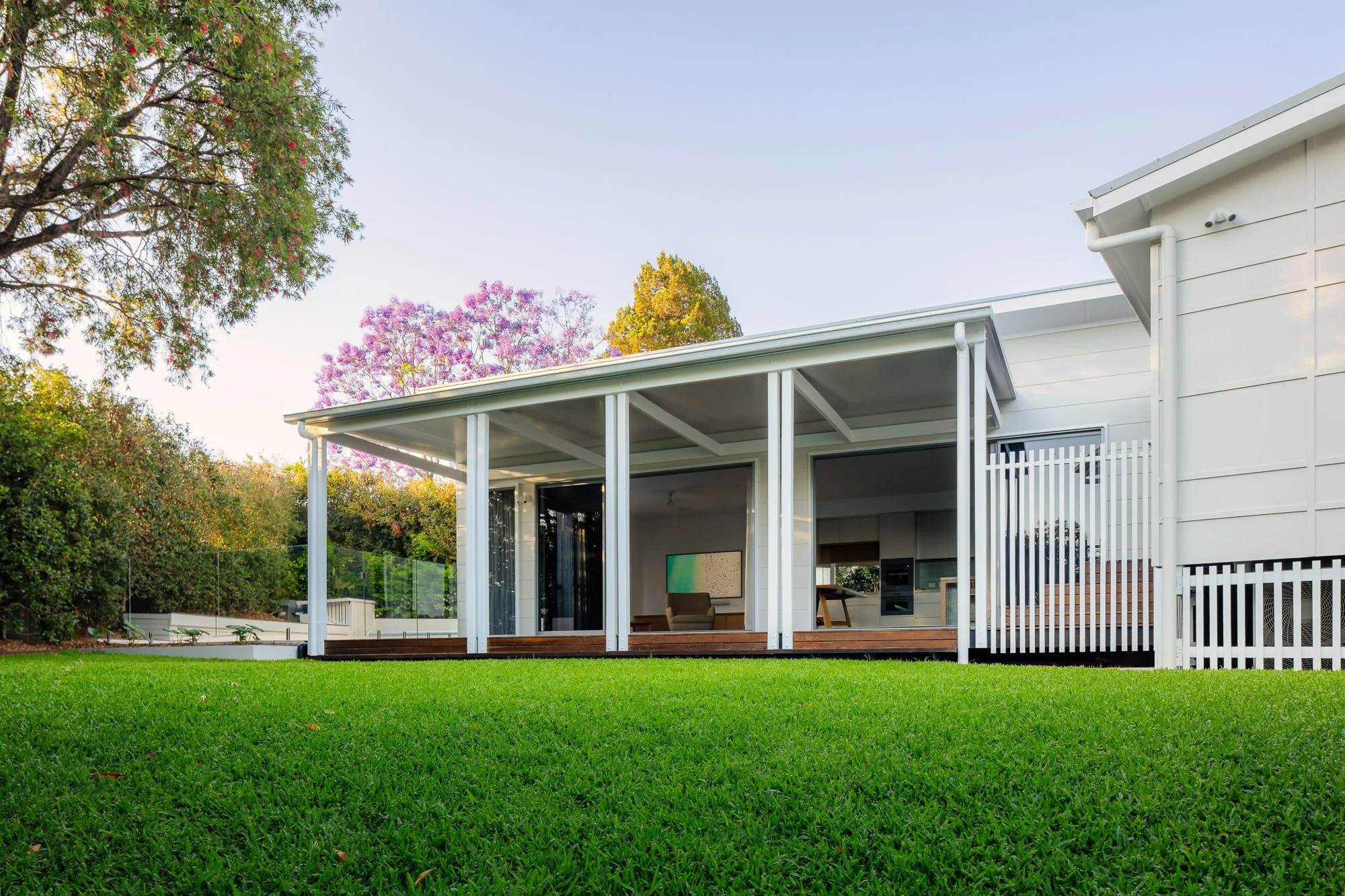 Modern house with white exterior, large front porch with seating, surrounded by green lawn and trees with pink and purple flowers, clear sky in the background.