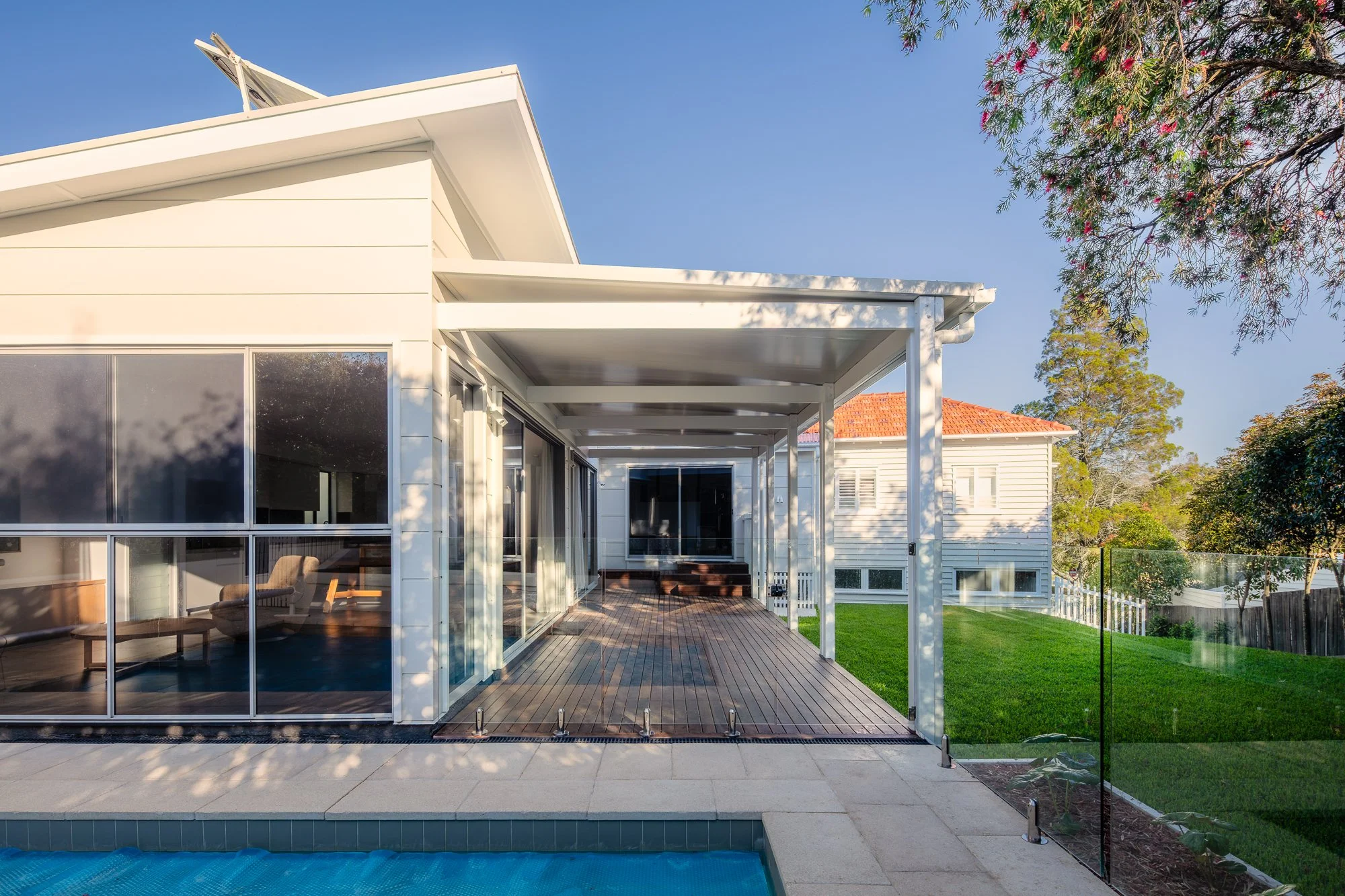 Modern house with a screened-in porch, wooden deck, and glass sliding doors. The house has white siding, a slanted roof with solar panels, and is situated in a green yard with trees and a clear blue sky.