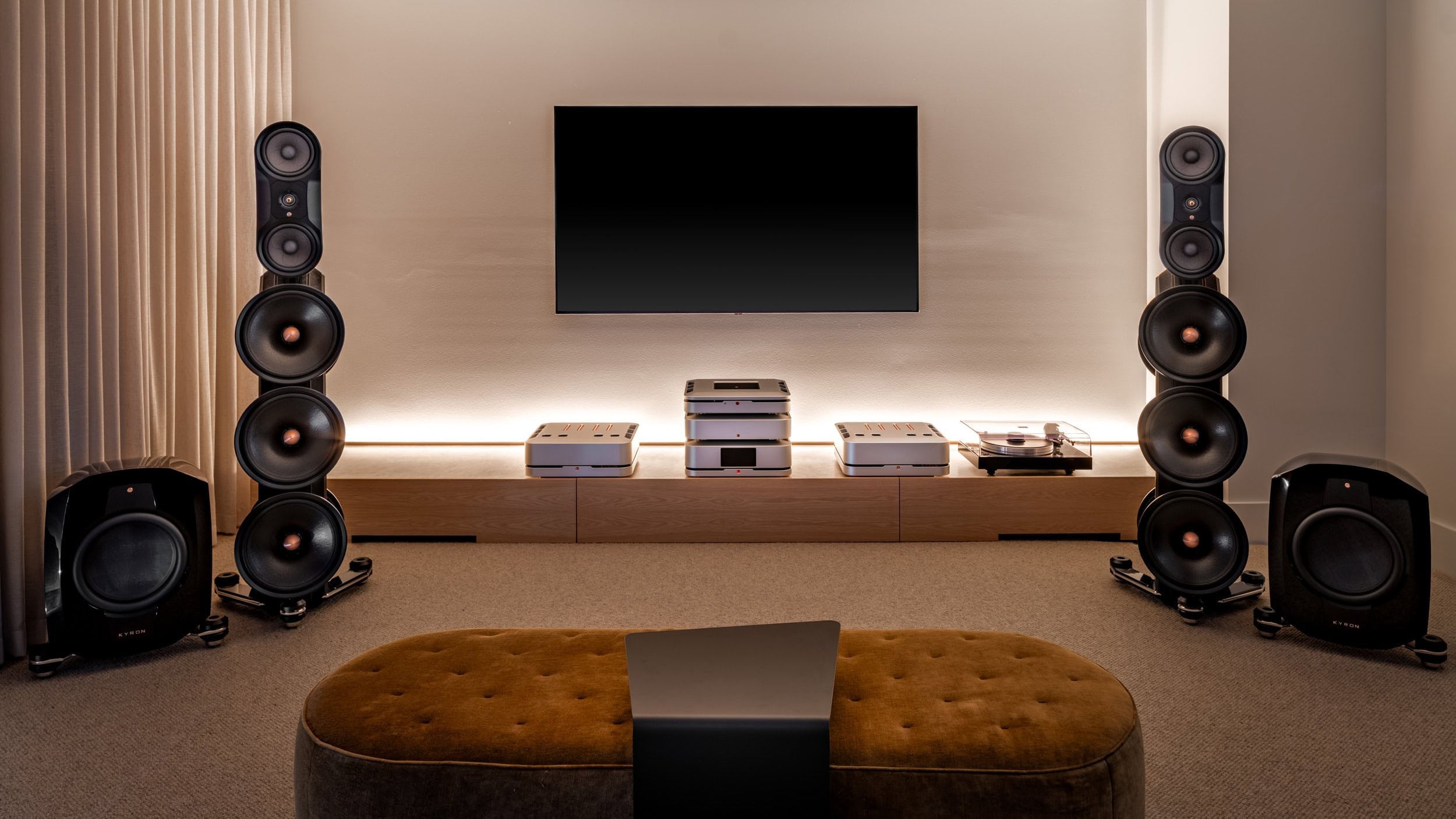 Modern home entertainment room with two large tower speakers by Kyron Audio, two large subwoofers, a flat-screen TV, audio equipment, a turntable, and an oval velvet ottoman