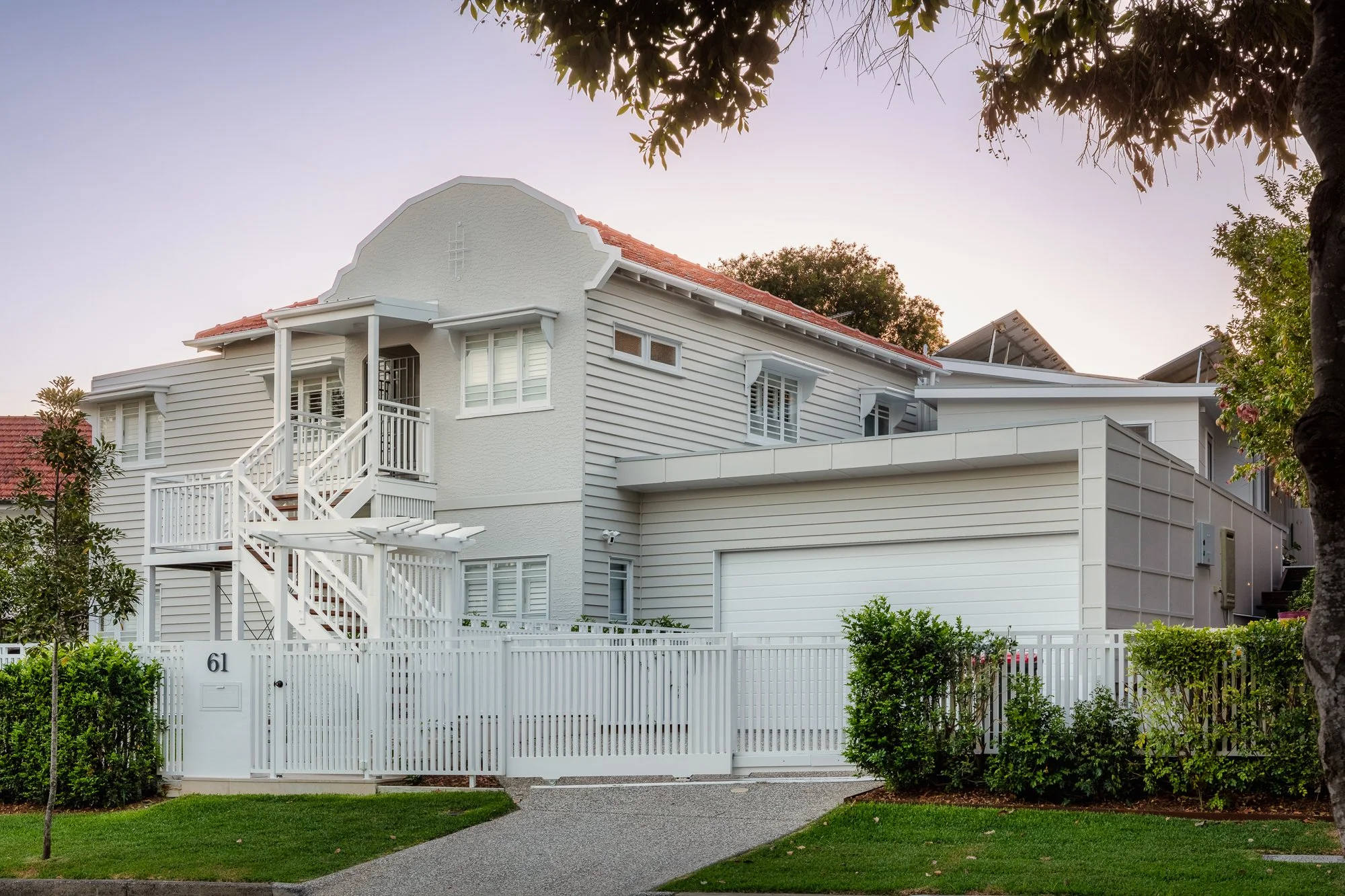 A white multi-story house with a red-tiled roof, surrounded by a white fence and lush green grass, with blue sky and trees in the background.