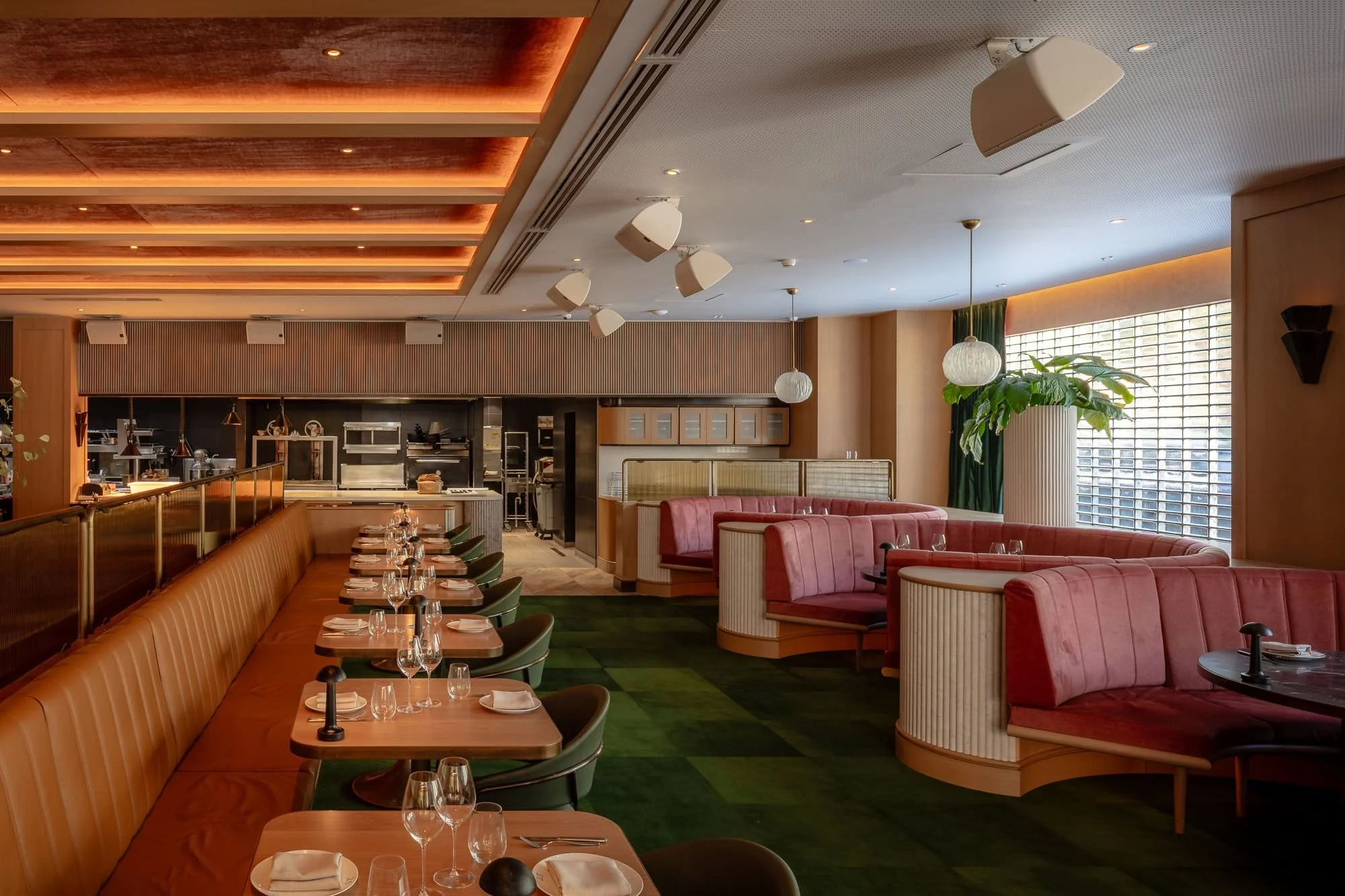 Interior of a restaurant with pink curved booths and green chairs, wooden tables set with glasses and napkins, large windows with blinds, ceiling lights, and kitchen area in the background.