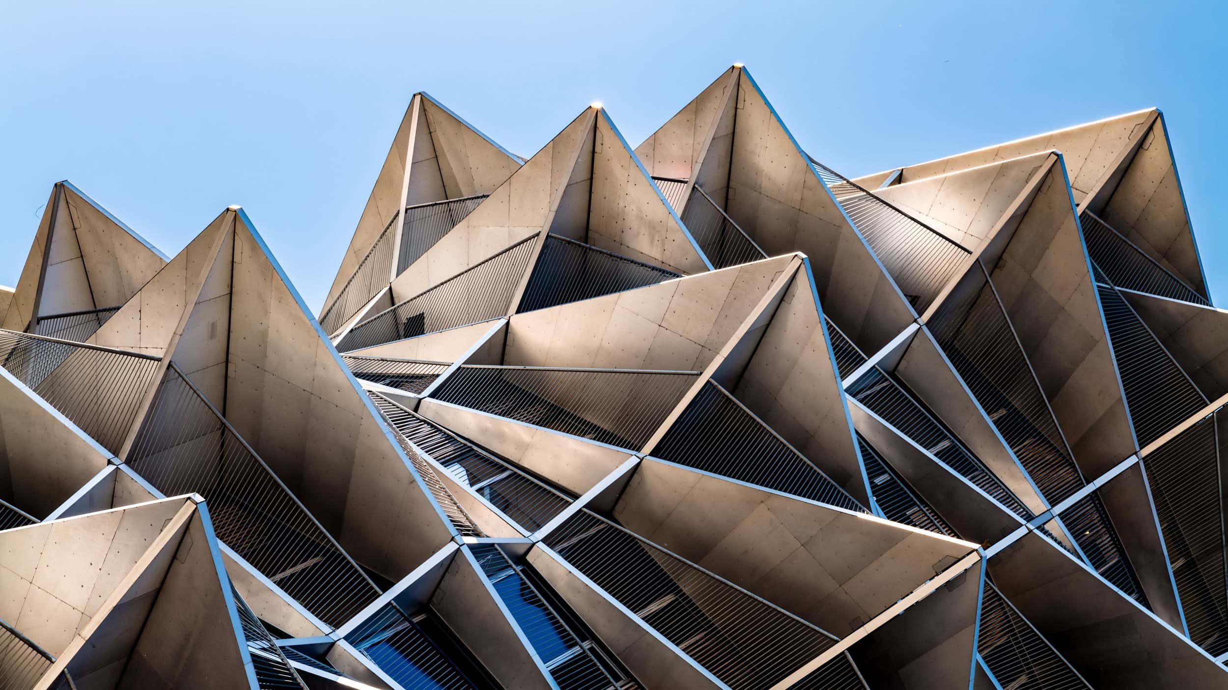 Modern building with angular concrete and glass exterior, featuring sharp peaks and multiple terraces, against a clear blue sky.