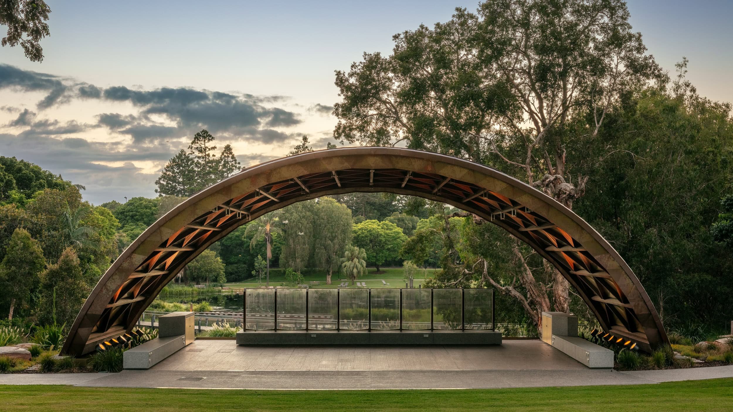 A modern amphitheater with a curved, semi-circular roof structure in a park setting, surrounded by trees and a lake with a cloudy sky in the background.
