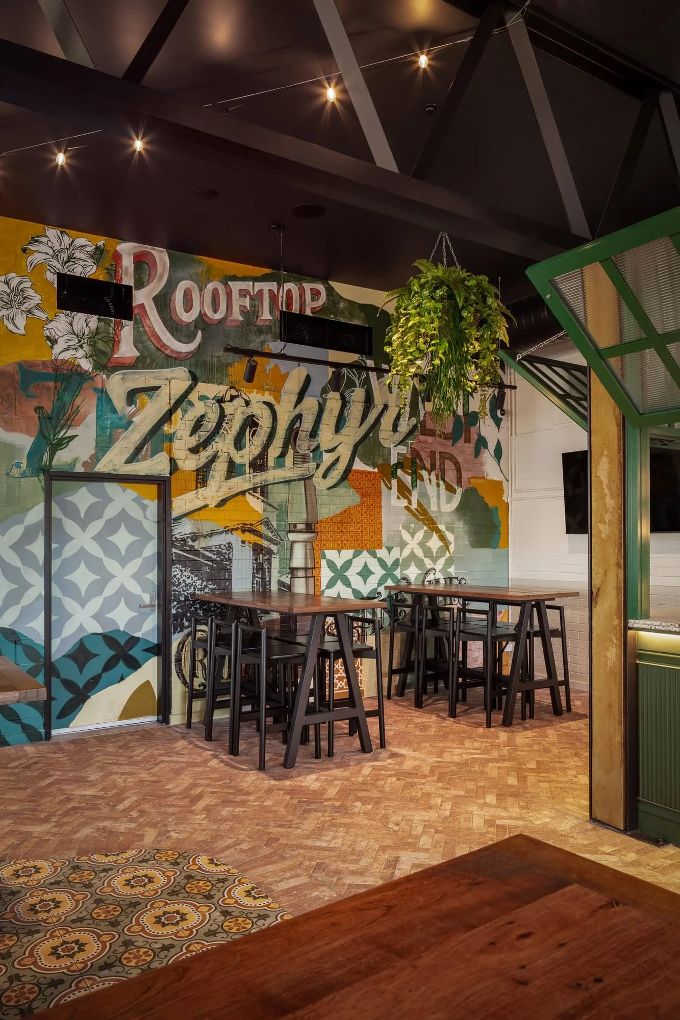 Interior of a modern restaurant with a colorful floral mural on the wall, high tables and black stools, hanging greenery, green framed window, black ceiling with lighting, and patterned tile and wood flooring.
