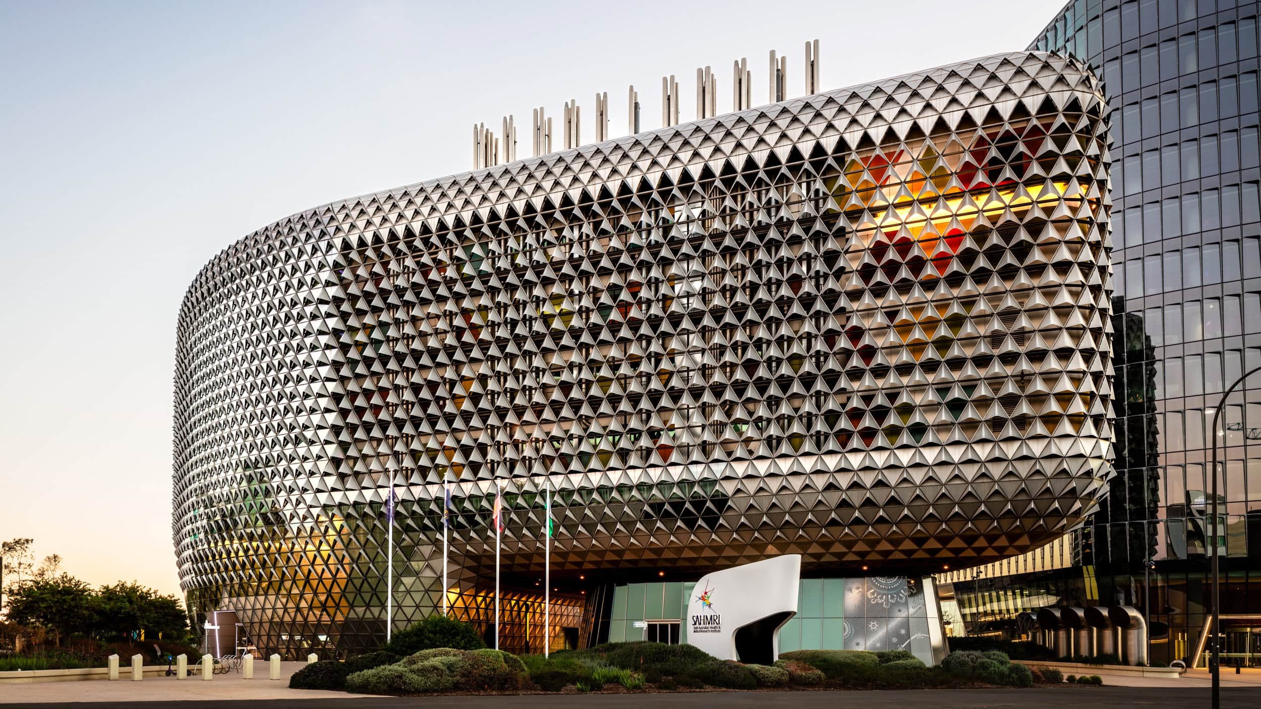 Modern building with a geometric facade, glass windows, flags, and a sign that reads 'SAHMRI' at sunset.