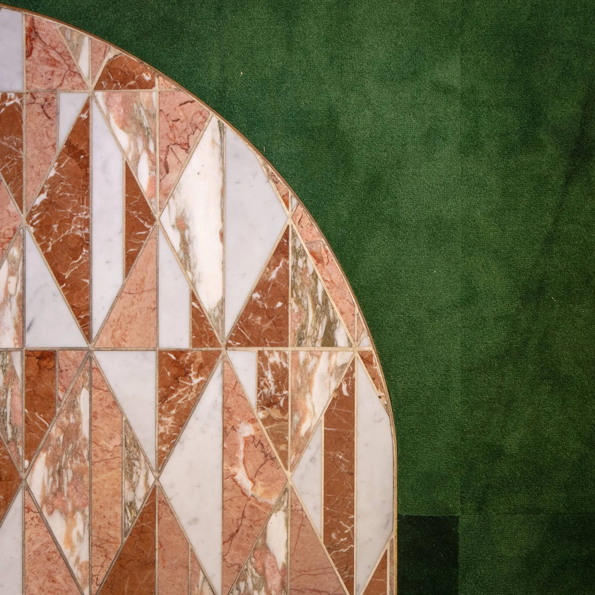 Close-up view of a semi-circular terrazzo table surface with a pattern of white, pink, and brown marble pieces, next to artificial green grass.