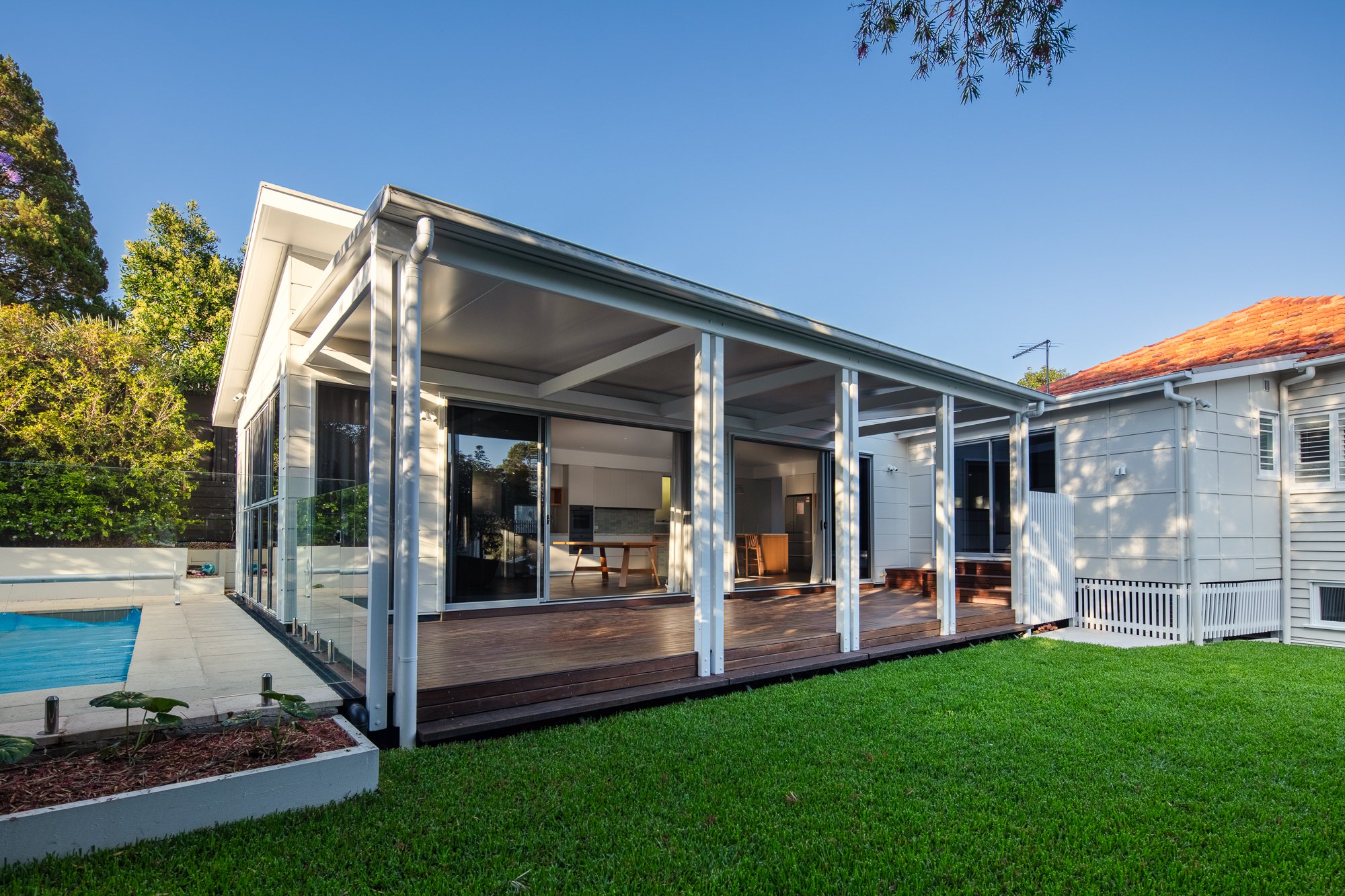 A modern backyard with a house featuring a glass-enclosed patio area with white framing, wooden decking, a swimming pool, and well-maintained green grass with trees surrounding the yard.