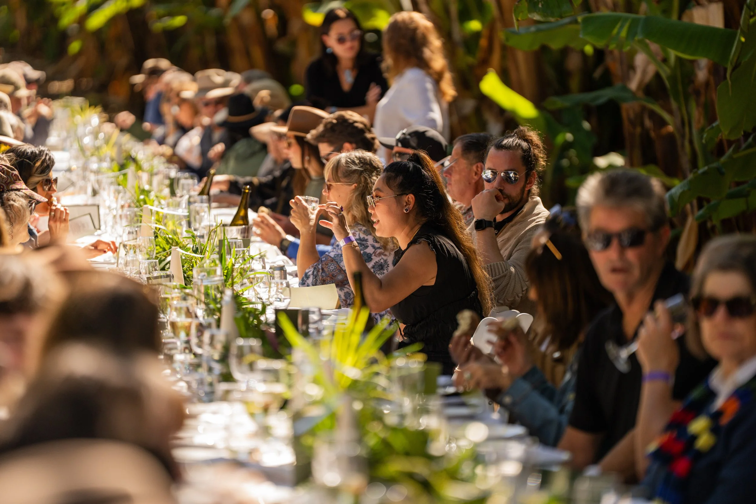 People attending an outdoor event seated at long tables with glassware, beverages, and greenery, enjoying daytime socializing in a lush environment.