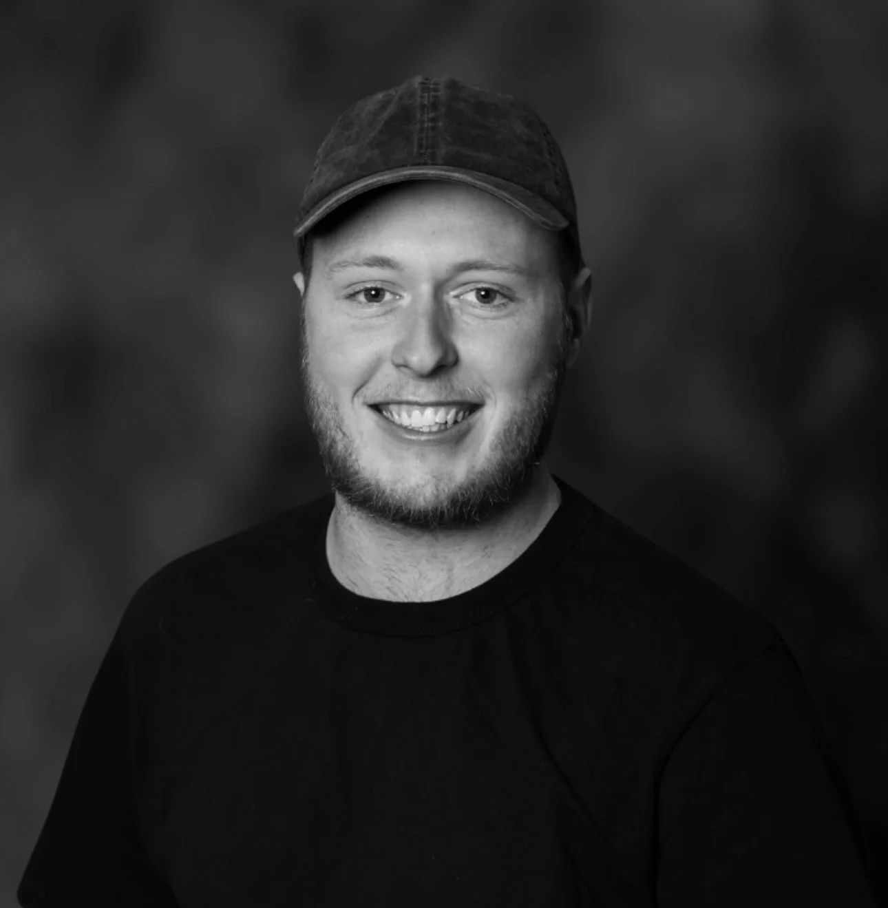 A smiling young man with a beard and wearing a cap and a black t-shirt, standing against a blurred background.