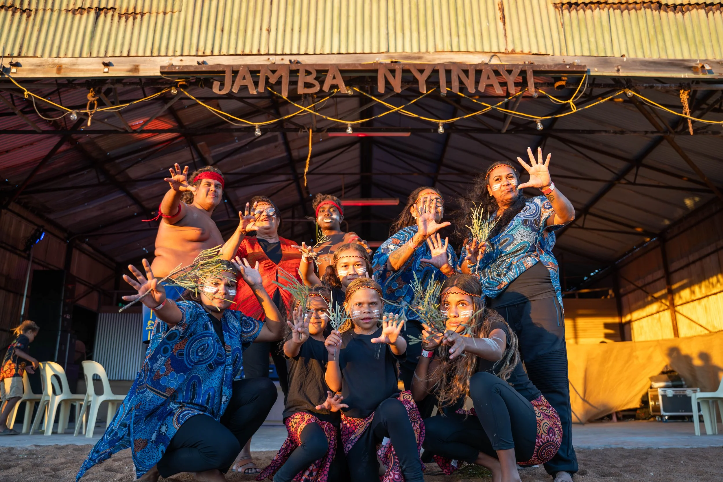 Group of people in colorful clothing performing a traditional dance on an outdoor stage with a sign above that reads 'JAMBA NYINAYI' at sunset.