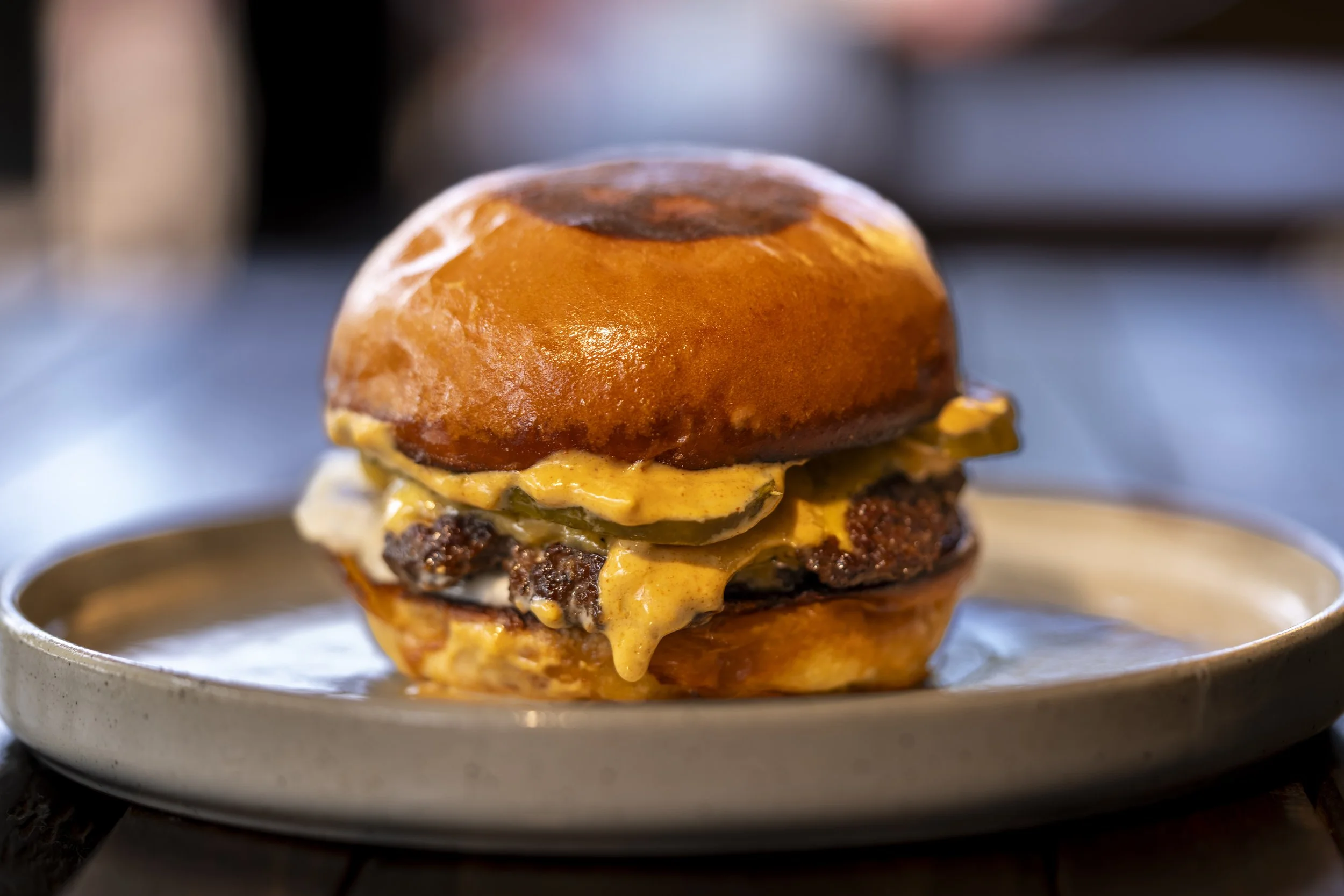 Close-up of a cheeseburger with a shiny brioche bun, melted cheese, pickles, and a beef patty on a plate.