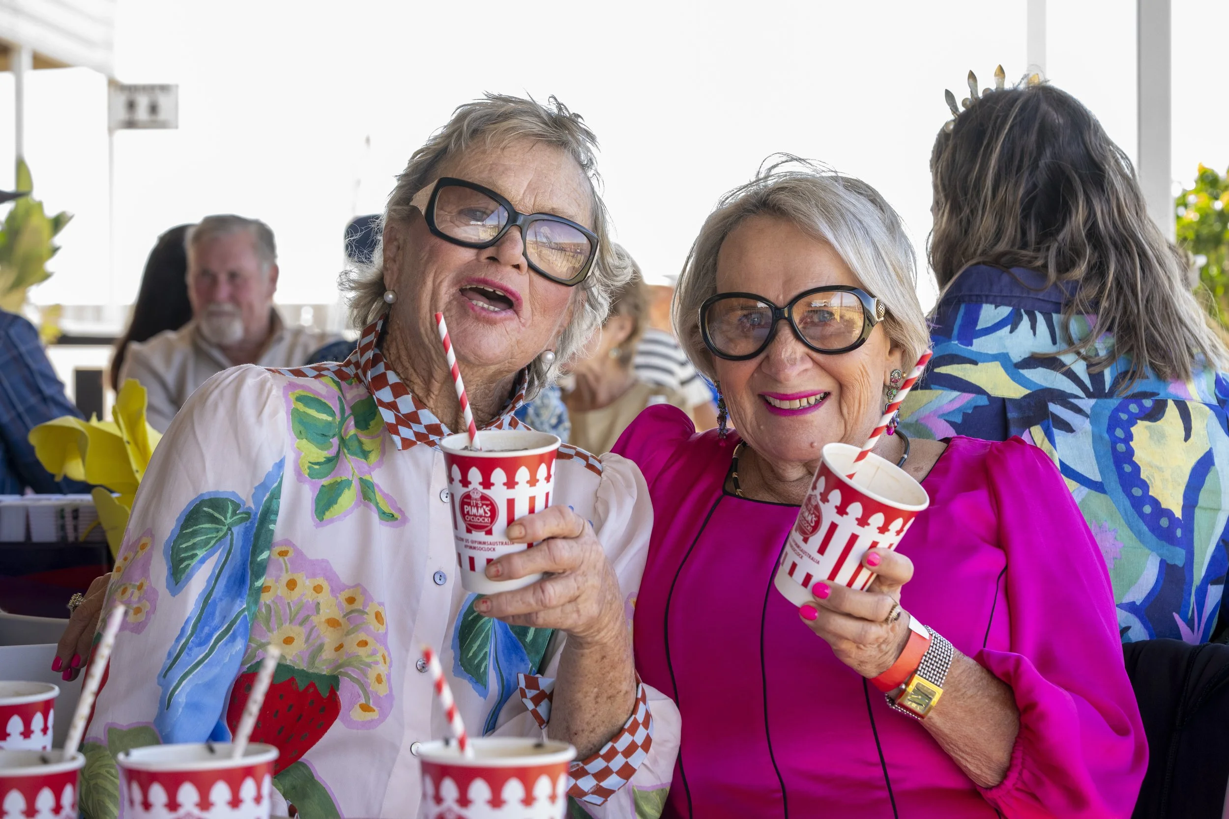 Two elderly women with glasses and colorful outfits enjoying drinks with striped straws at a social gathering, smiling at the camera.
