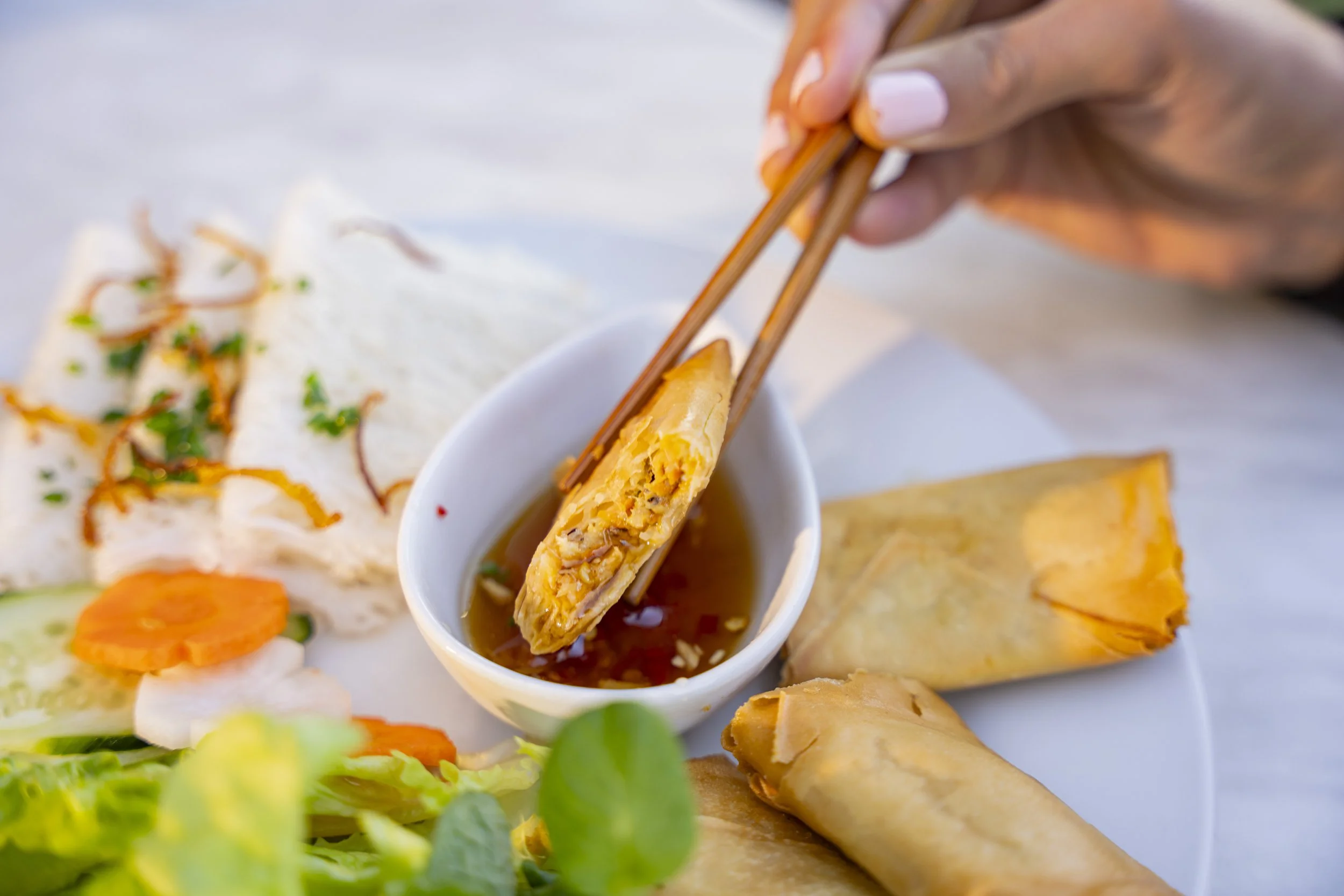 Person using chopsticks to dip a spring roll into dipping sauce on a plate with additional spring rolls, sliced vegetables, and salad.