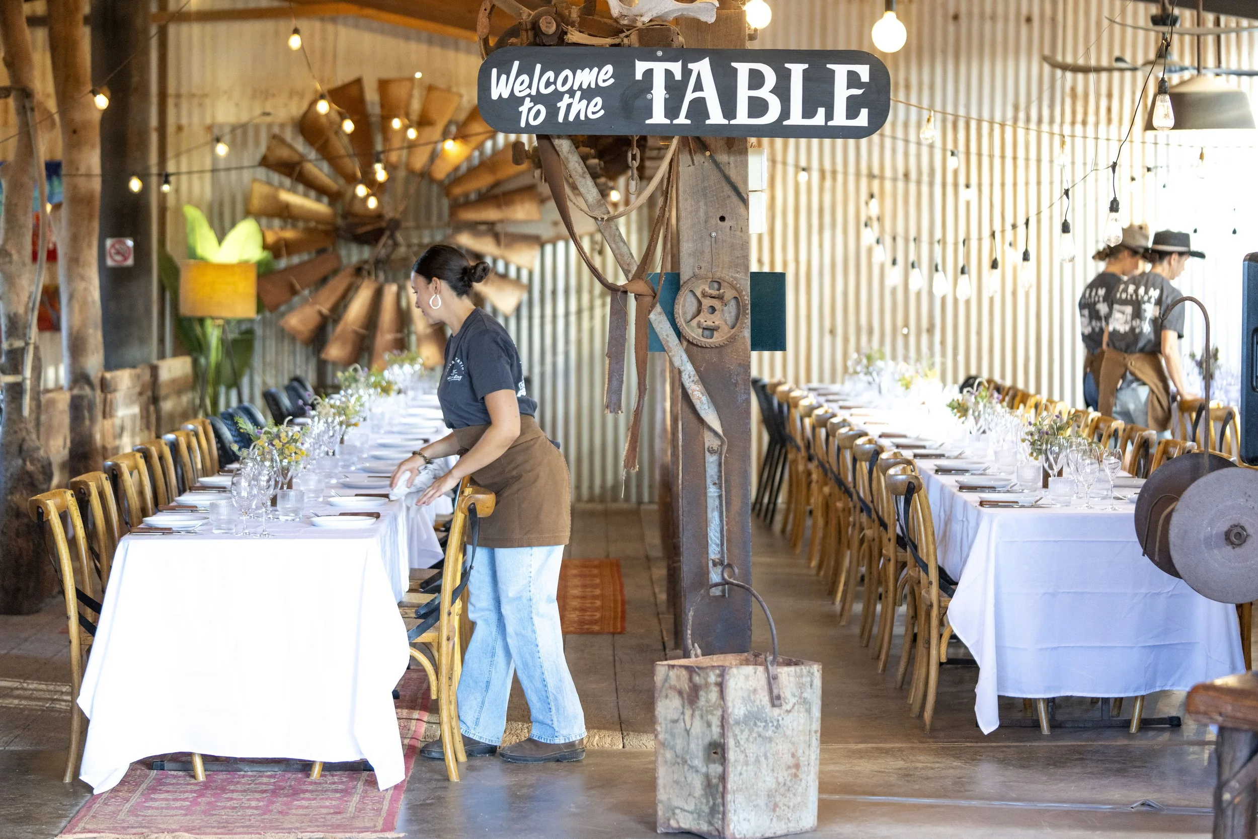 A rustic restaurant interior with long tables covered in white tablecloths, set for a meal with glasses and cutlery, person arranging the table, and a sign that says "Welcome to the TABLE" hanging from a wooden post, with warm lighting and decorative