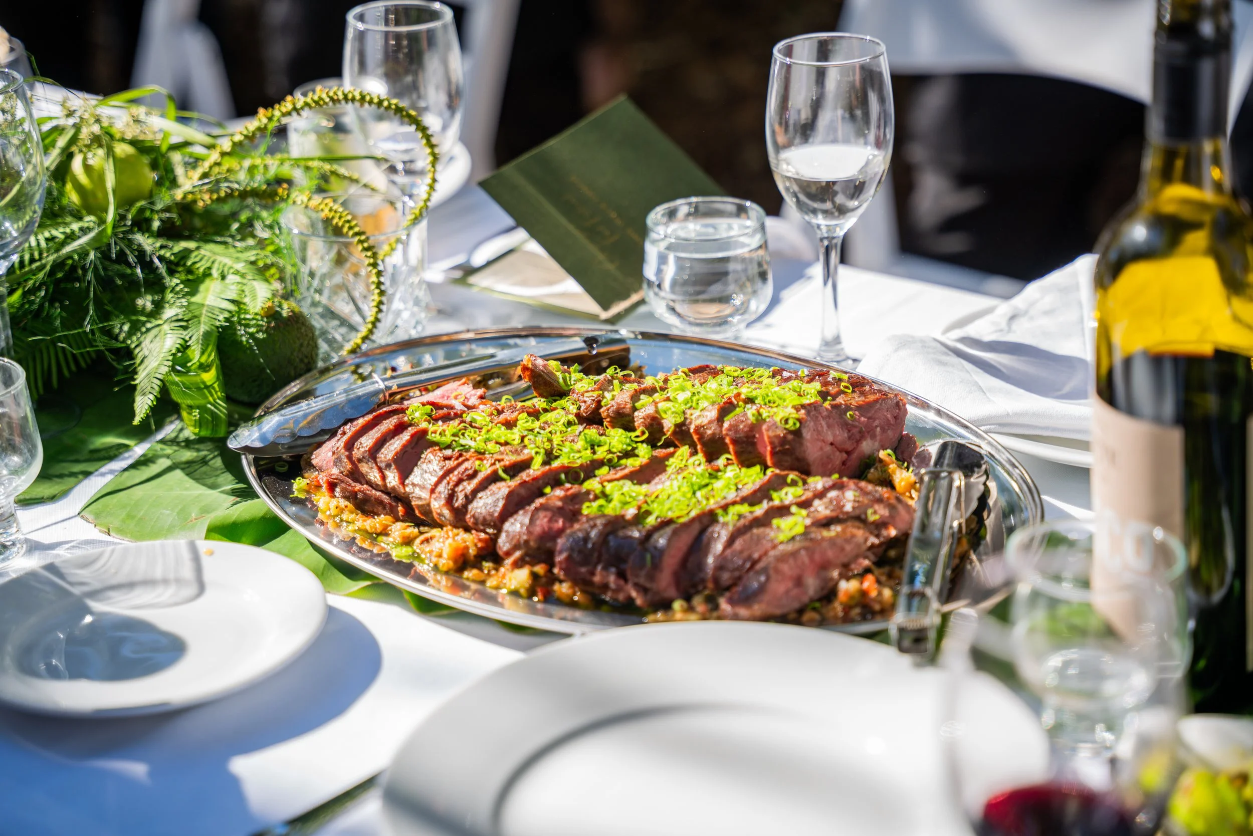 Sliced cooked beef garnished with green onions on a silver platter at a formal dining table with glasses, a green leafy centerpiece, and a bottle of wine.