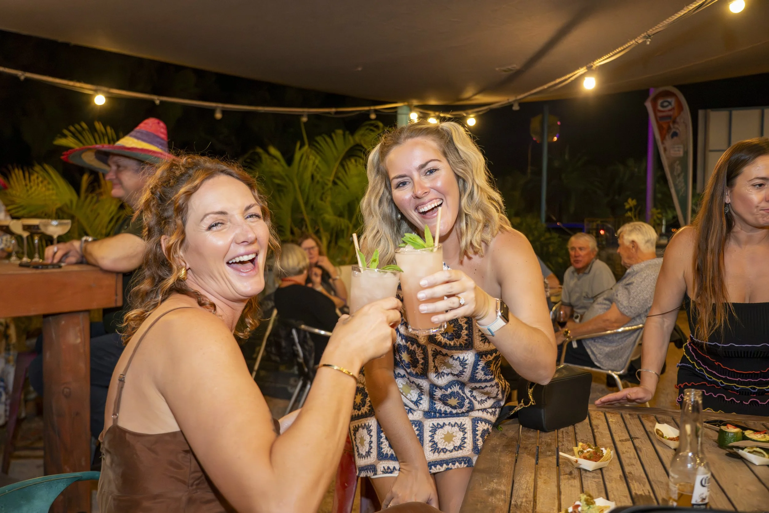 Two women are smiling and raising cocktails at a nighttime outdoor gathering, celebrating and enjoying drinks.
