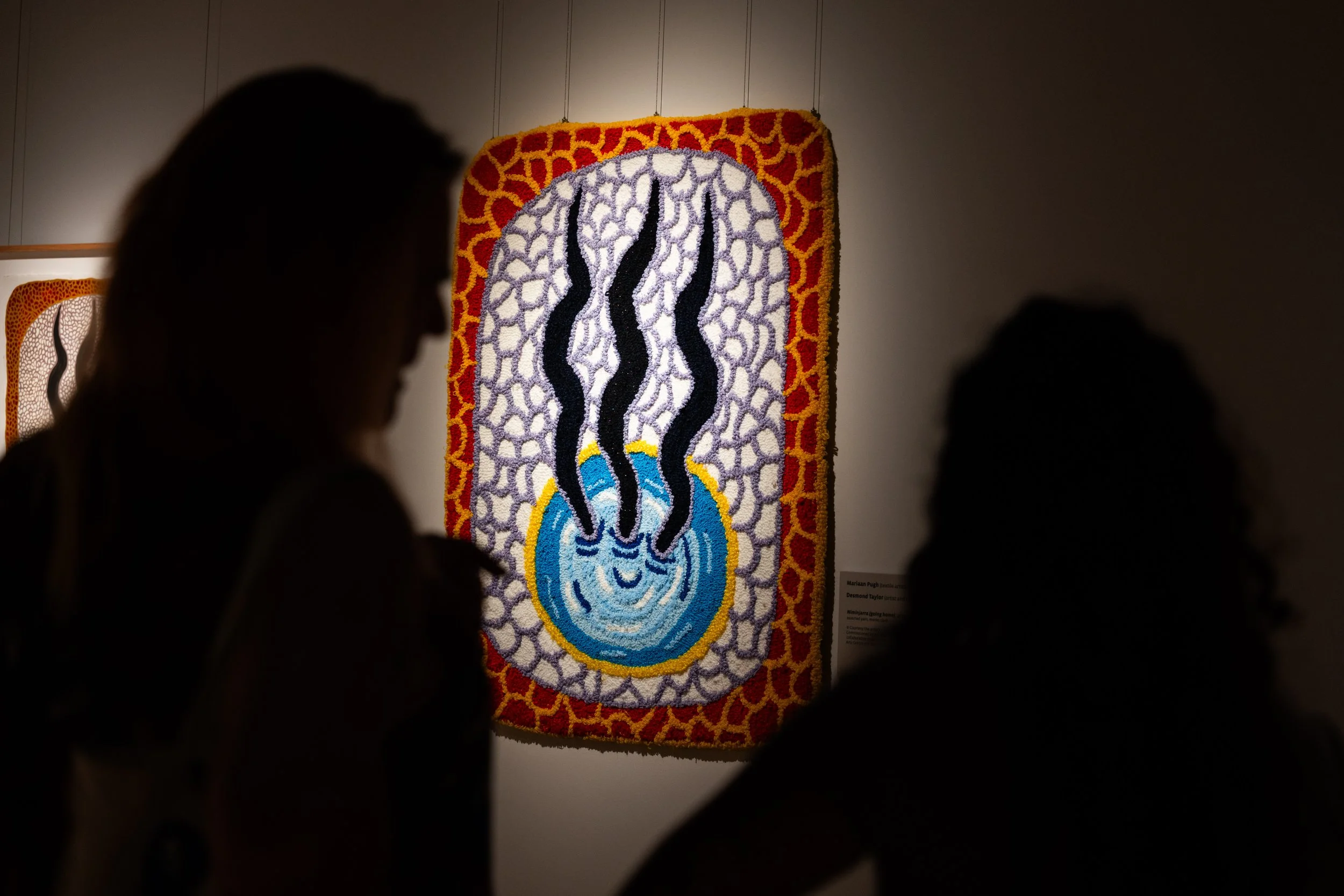 Silhouettes of two women looking at a colorful, textured artwork featuring three black wavy lines rising from a blue circle with a yellow edge, on a white background with a red border.