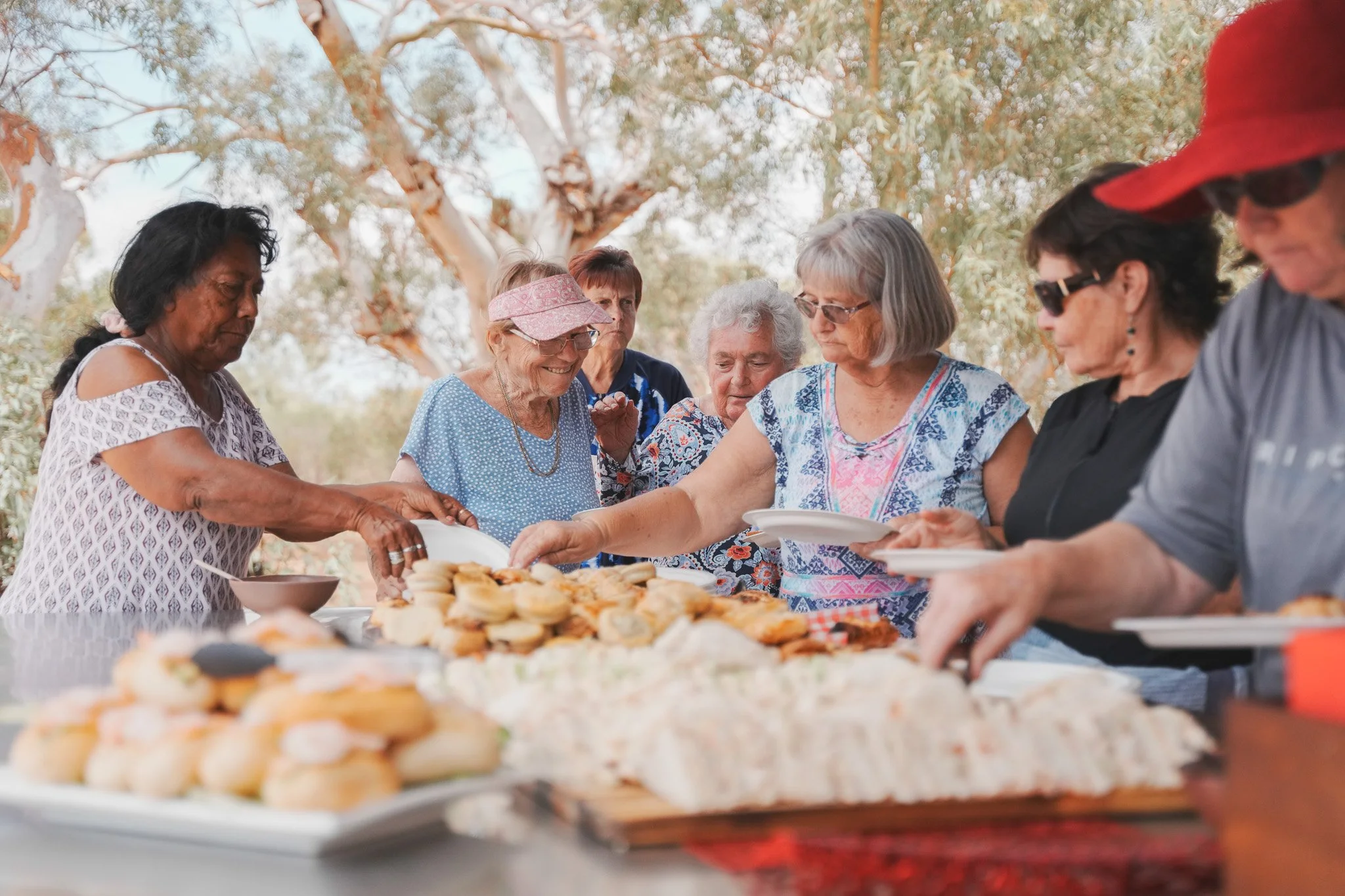 A group of elderly women and one man gathering outdoors around a table with a variety of pastries and desserts, serving themselves from the spread during a social event or gathering.