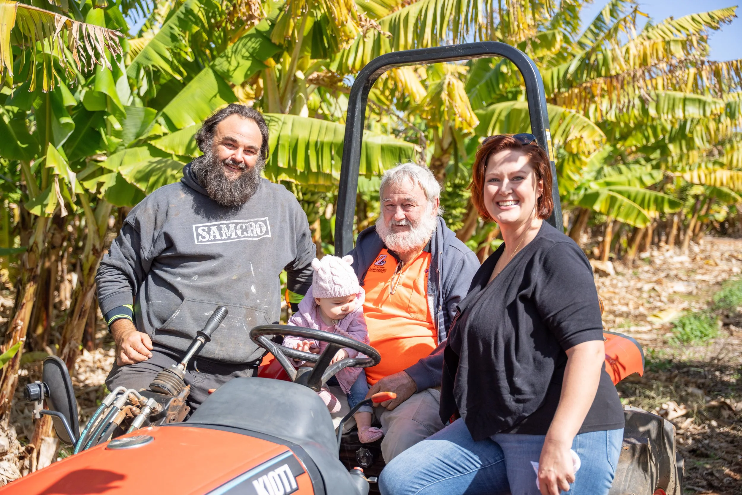 A group of four adults and one child sitting on and standing near a tractor in a banana plantation, smiling at the camera.