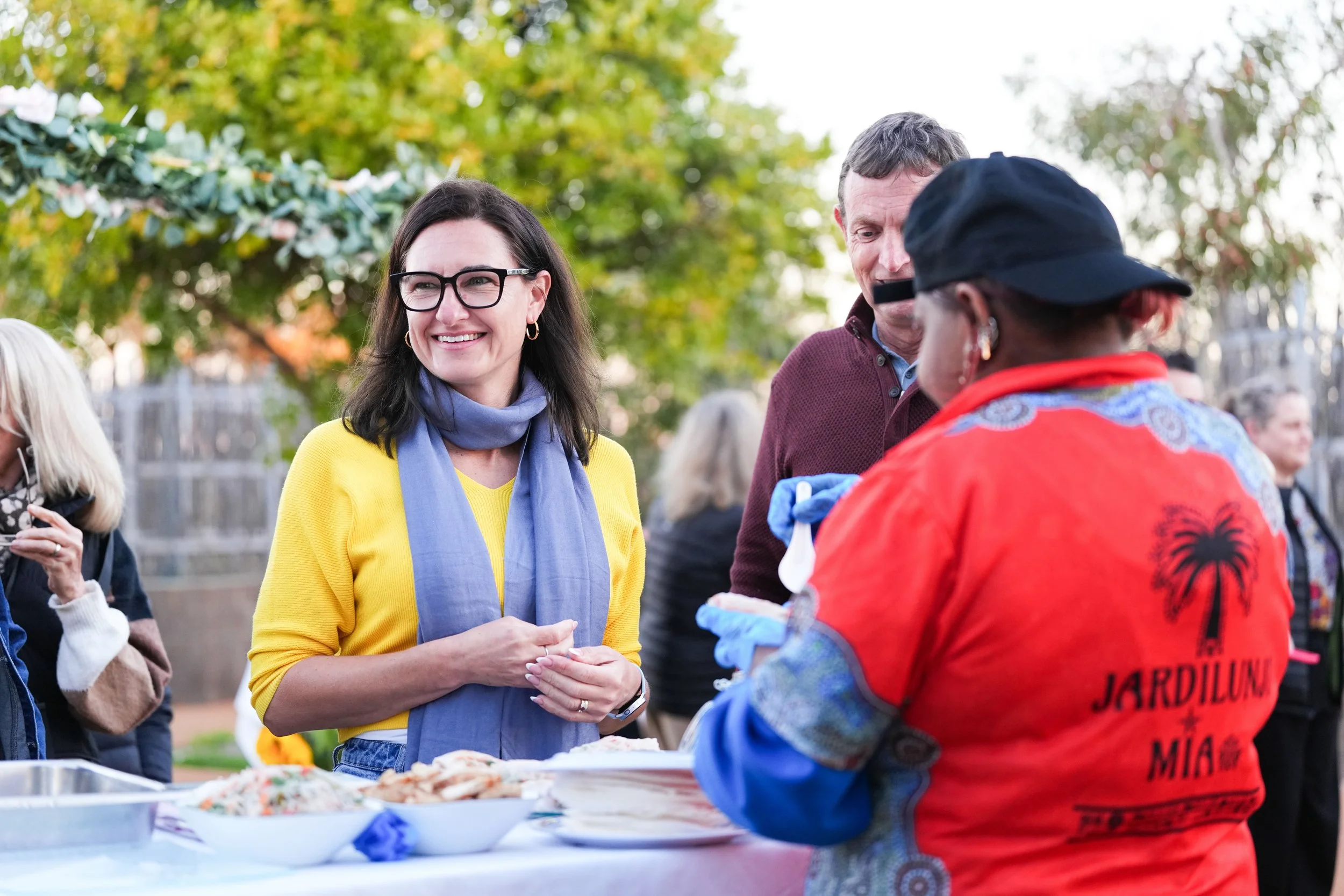 A woman in a yellow shirt and glasses smiling at a food stand outdoors, talking with a woman in a red shirt and black cap, with a man standing nearby, in a park setting with trees in the background.