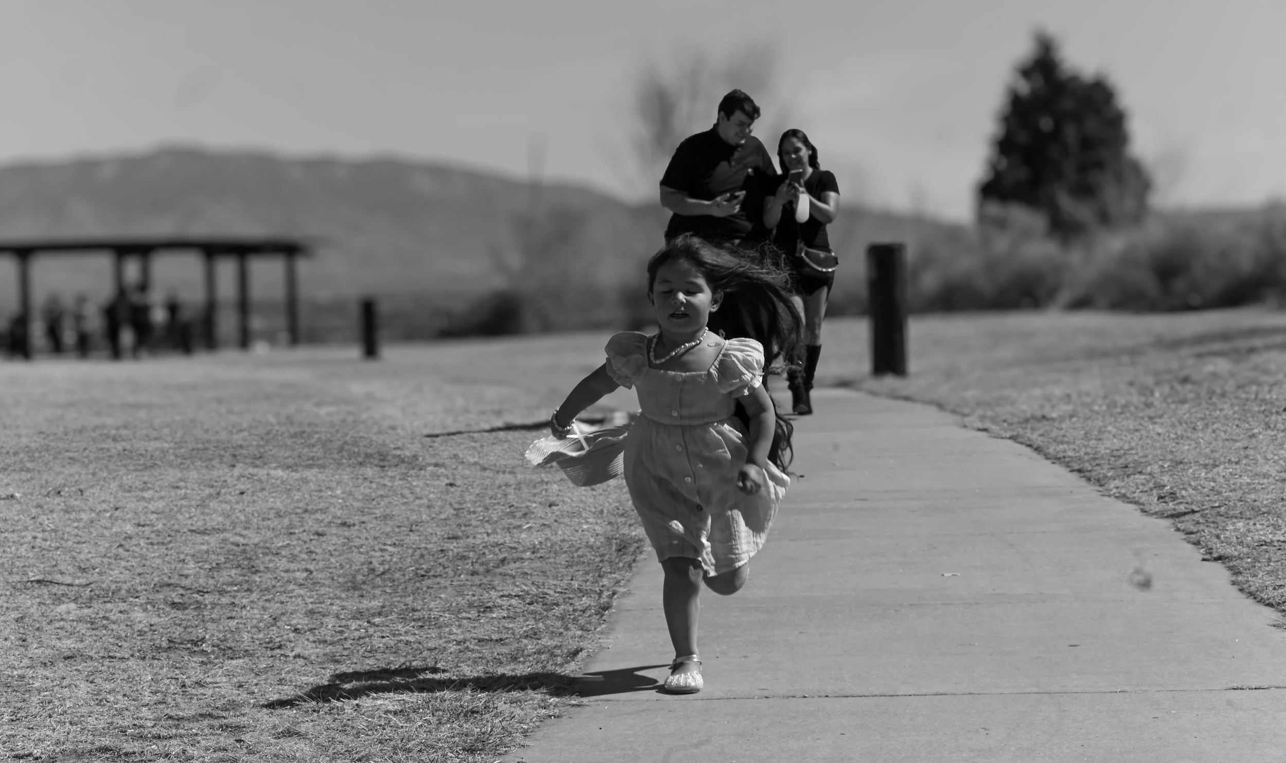 A young girl running on a sidewalk with wind-blown hair, wearing a dress and sandals, with a group of people including a man and woman looking at their phones in the background, outdoors in a rural area.