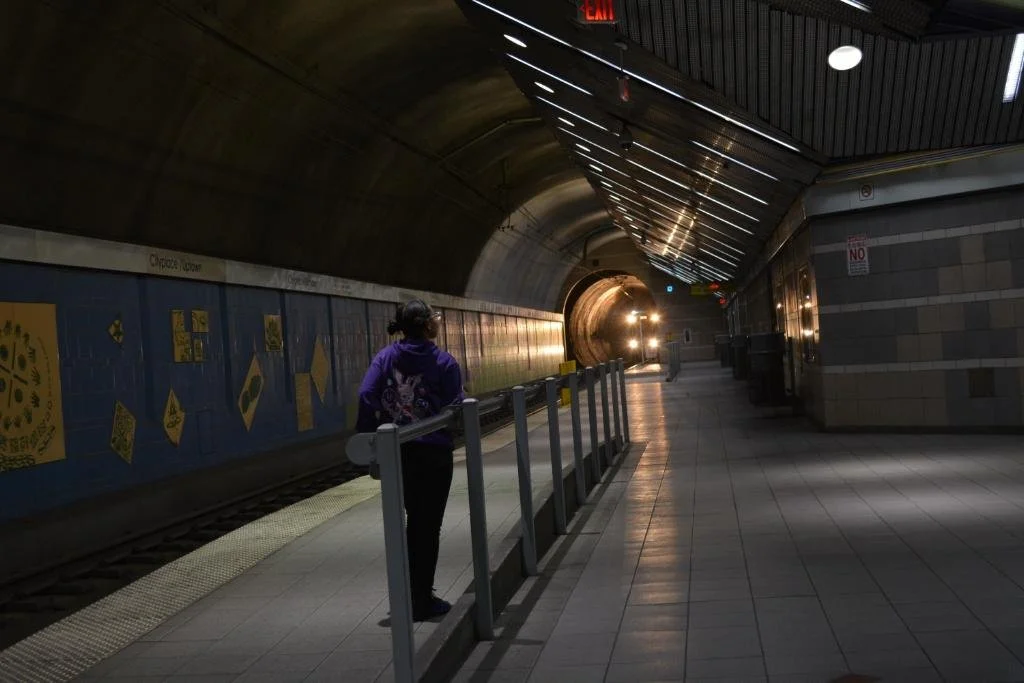 A woman in a purple hoodie waiting on a subway platform with a train approaching in a tunnel.