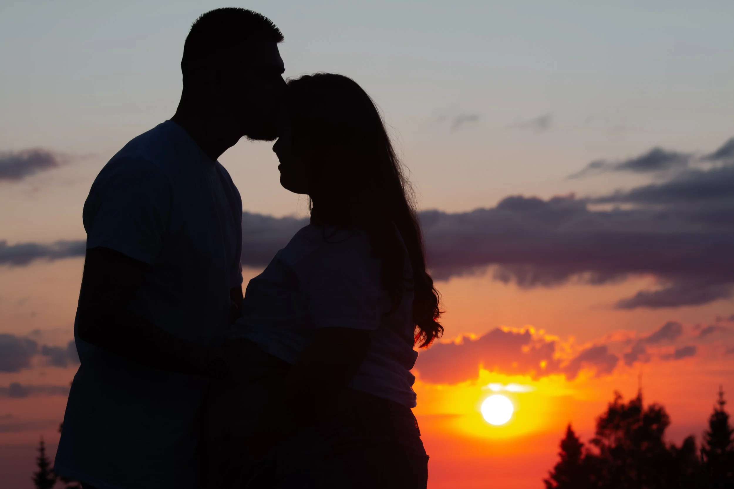 Silhouette of a couple kissing at sunset with an orange sky and trees in the background.