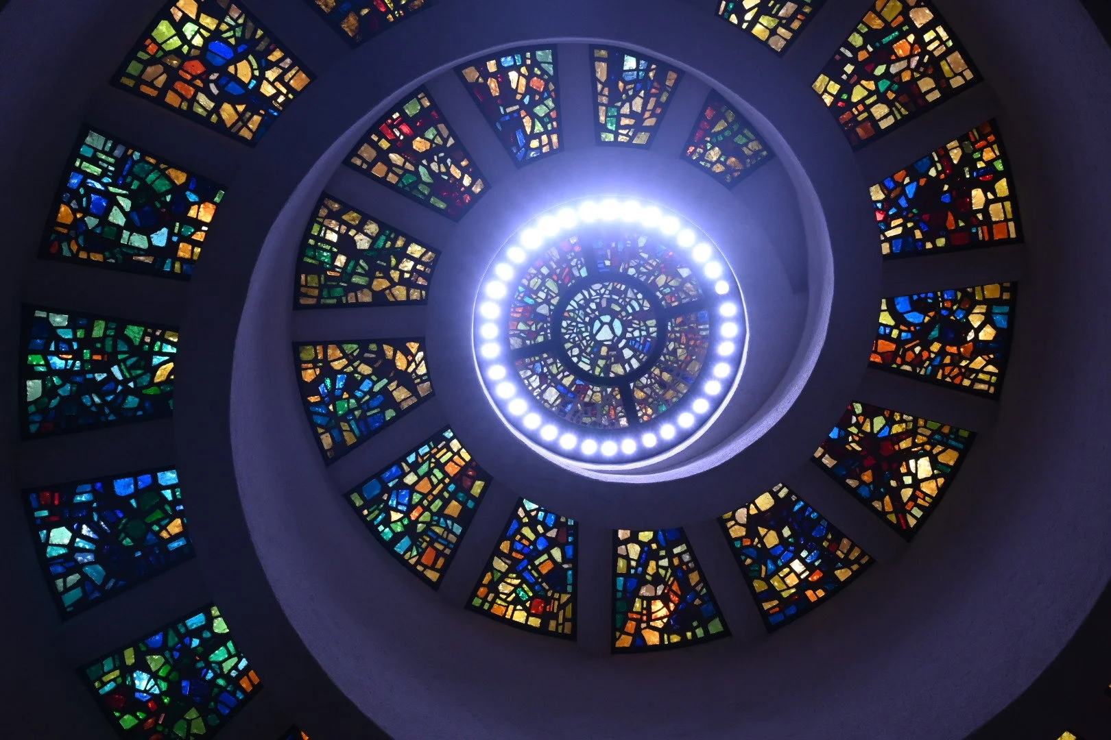 Looking up at a spiral staircase with colorful stained glass windows and circular lights at the center.