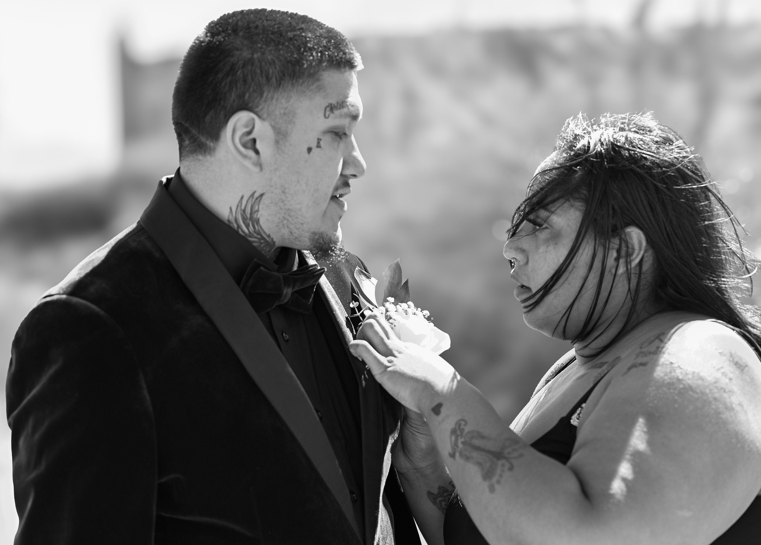 A groom and bride stand close together in a wedding ceremony outdoors, facing each other, with the groom wearing a tuxedo and the bride with wild hair, holding a flower, concentrating on each other.