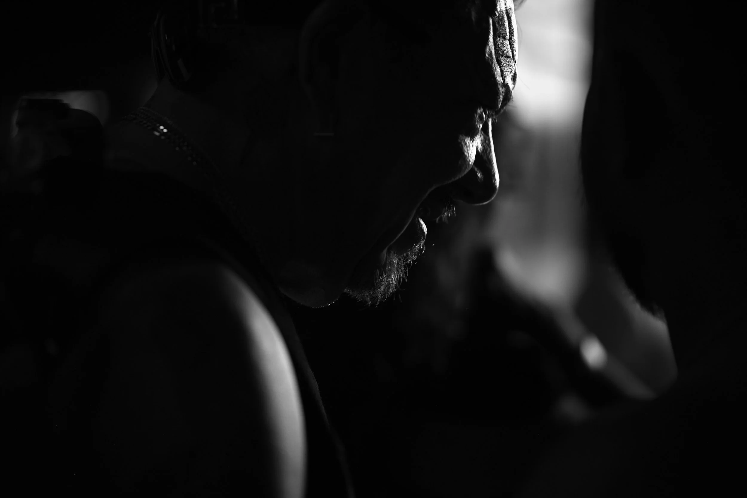 A close-up profile of a person's face and shoulders in black and white, with dramatic lighting highlighting facial features and textured skin.