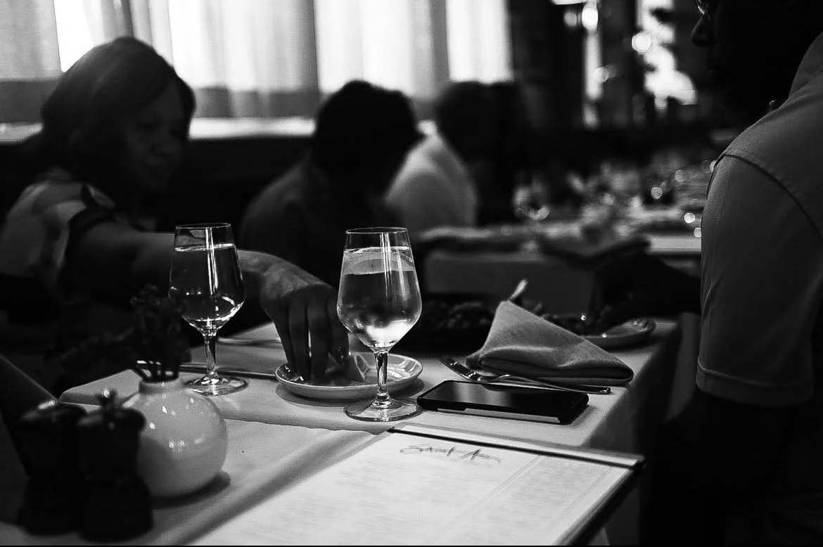 People seated at a restaurant table, with water glasses and place settings, during a dimly lit dining scene.