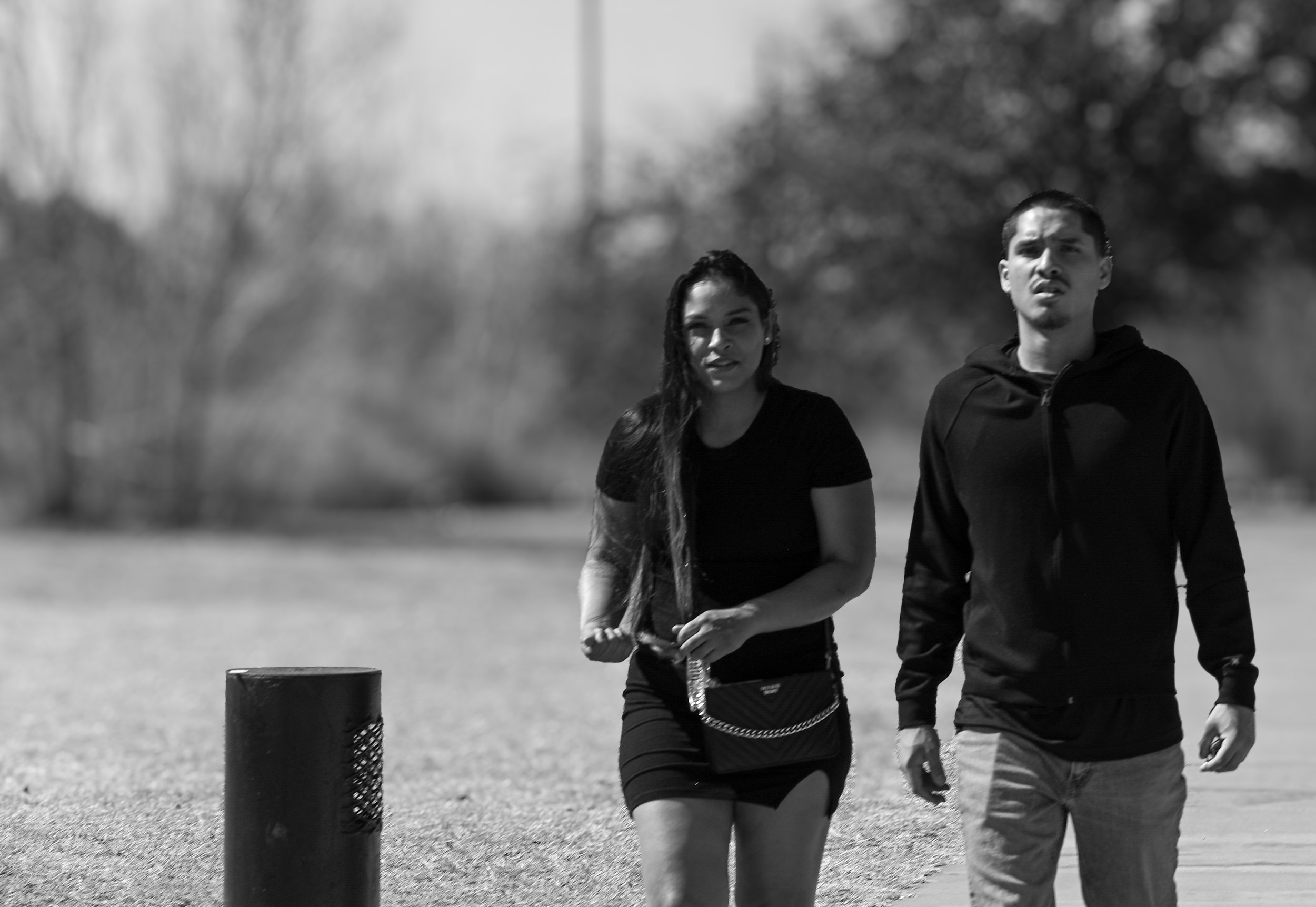 A black and white photo of a young woman and man walking outdoors on a sunny day. The woman has long hair, and the man has short hair. They are dressed in casual sportswear, with trees and a post visible in the background.